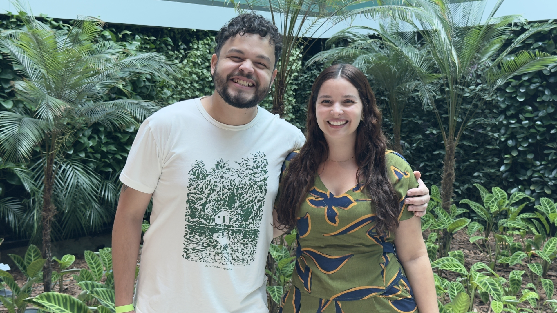 Duas pessoas posam sorrindo diante de um jardim interno com muitas plantas tropicais e folhagens verdes. À esquerda, um homem de pele clara, cabelo curto e cacheado e barba usa camiseta branca com estampa verde e coloca o braço em volta da mulher. À direita, uma mulher de pele clara e cabelos longos castanhos usa um macacão verde com estampas em azul e amarelo. Ambos olham para a câmera e aparentam descontração.