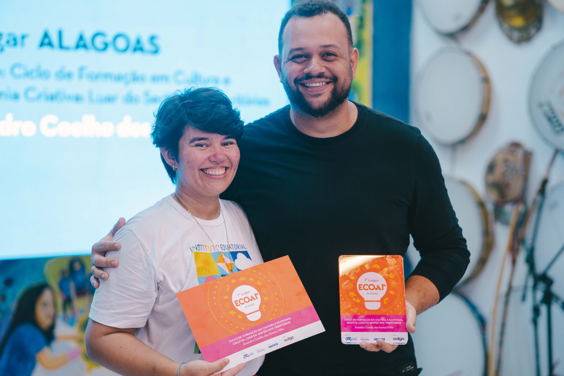 A imagem retrata dois participantes sorrindo durante a cerimônia do ECOAR Alagoas. Eles exibem orgulhosamente o certificado e o troféu do projeto premiado, em um ambiente marcado por iluminação suave e decoração cultural. O enquadramento aproxima os participantes, destacando a emoção e a celebração da conquista.