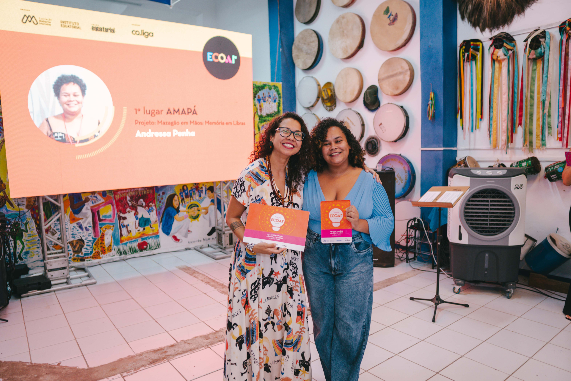 Duas mulheres sorriem para a câmera segurando um certificado e um troféu em um espaço cultural. À esquerda, uma mulher de vestido estampado e óculos abraça a participante ao lado. À direita, uma mulher de blusa azul clara e jeans segura o prêmio. No telão ao fundo aparece a inscrição “1º lugar Amapá” e o nome do projeto premiado. A parede ao lado é decorada com tambores e fitas coloridas penduradas.
