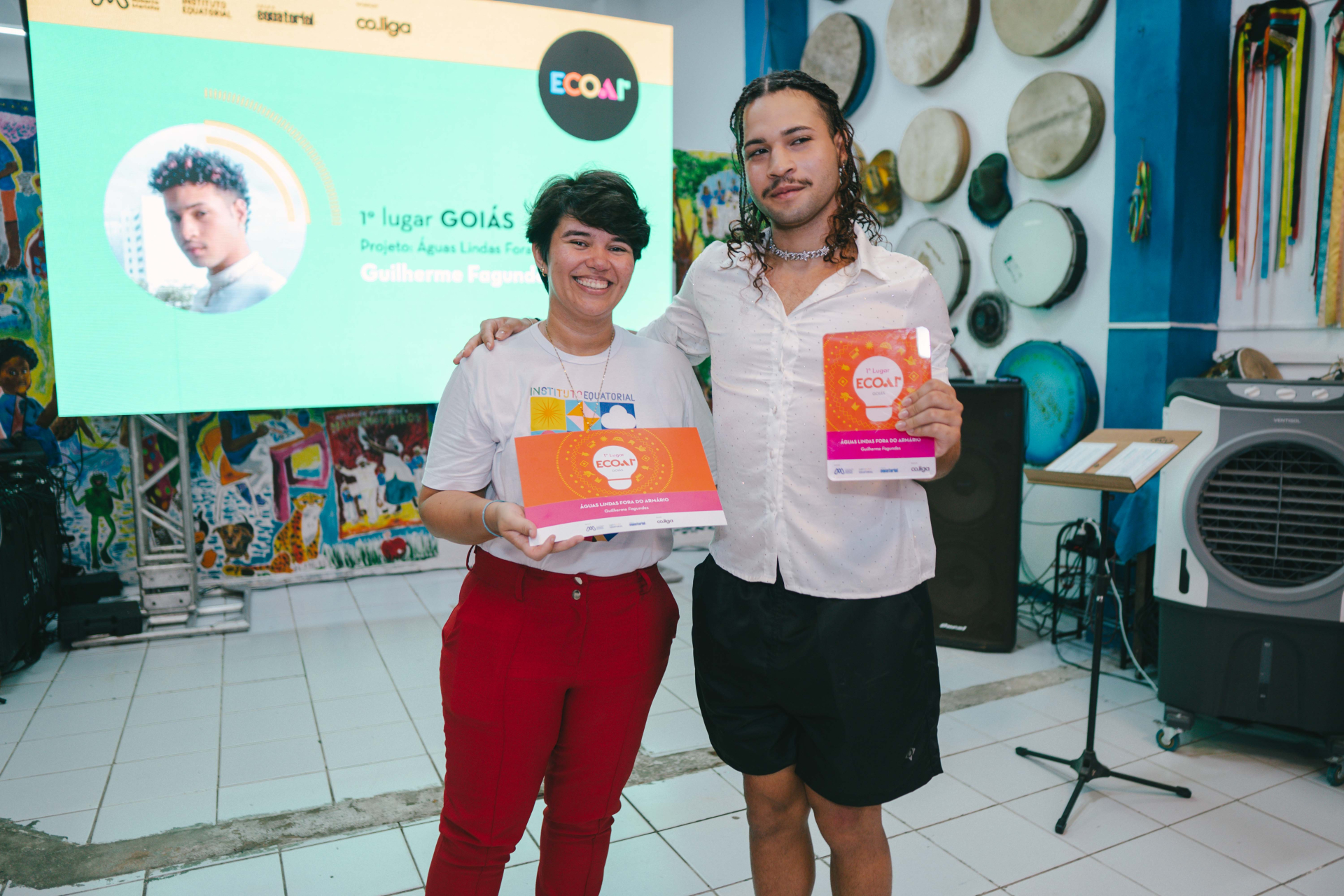 A fotografia registra a premiação de um participante de Goiás. Duas pessoas aparecem lado a lado segurando o certificado e o troféu do projeto vencedor. O telão ao fundo destaca o estado e o nome do premiado, enquanto os elementos decorativos — instrumentos musicais e obras artísticas — reforçam o caráter cultural do evento.
