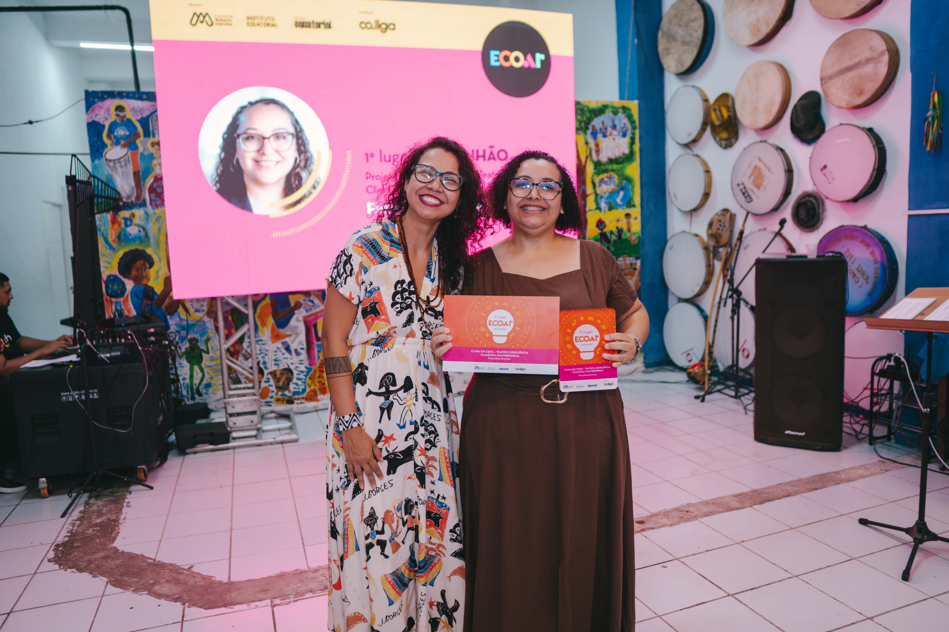 Duas mulheres posam sorrindo segurando o certificado e o troféu de 1º lugar do Maranhão. O ambiente é decorado com tambores e elementos artísticos, enquanto um grande painel iluminado ao fundo exibe a marca ECOAR e a foto da vencedora. A composição transmite um clima de celebração, reconhecimento e valorização cultural.