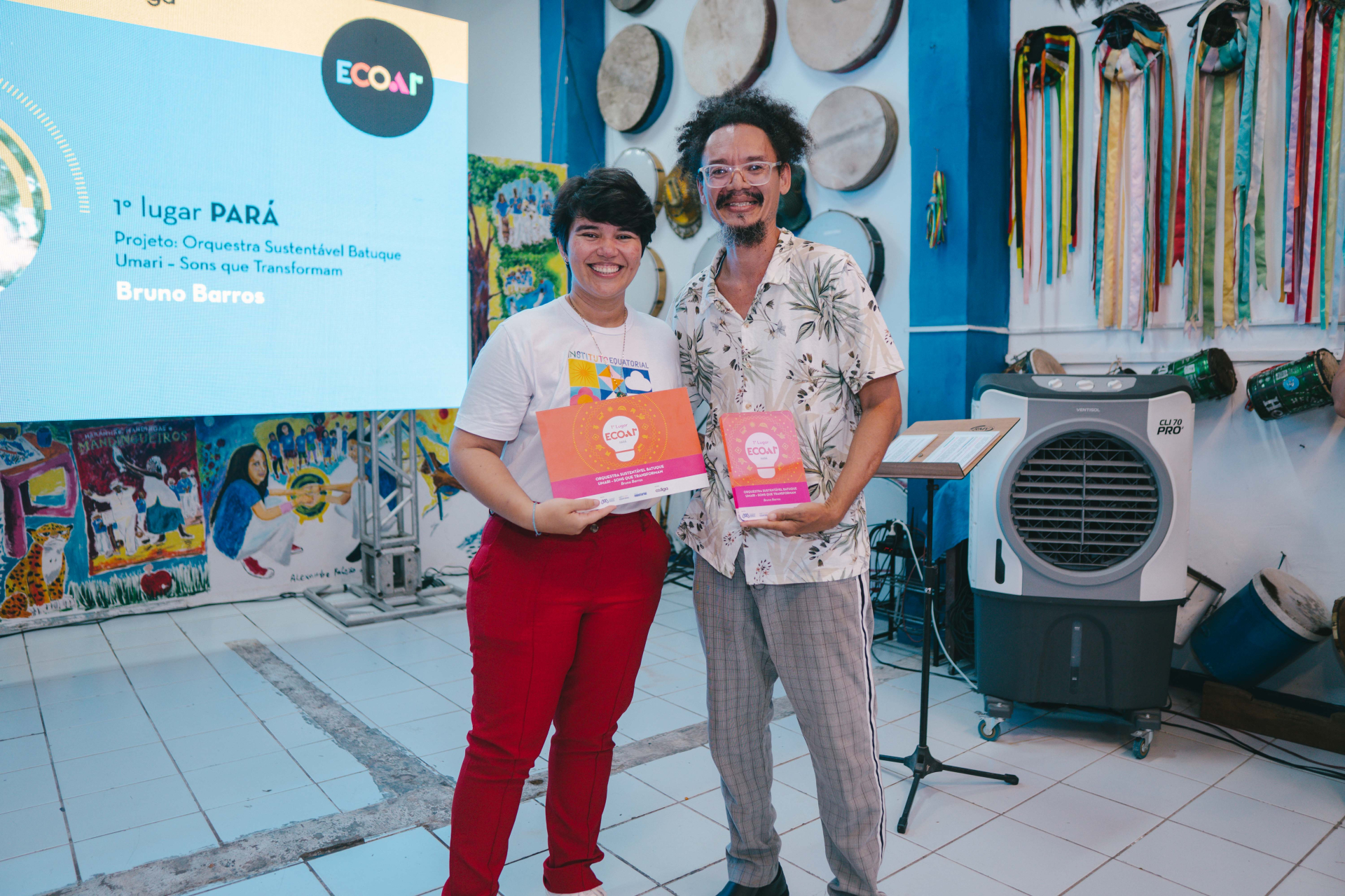 A imagem mostra duas pessoas sorrindo durante uma cerimônia de premiação do ECOAR. Elas seguram um certificado e um troféu referentes ao 1º lugar do Pará, com o projeto “Orquestra Sustentável Batuque Umari – Sons que Transformam”, de Bruno Barros. Ao fundo, há um telão com a identidade visual do prêmio, além de instrumentos musicais pendurados na parede e pinturas coloridas que remetem à cultura popular brasileira.