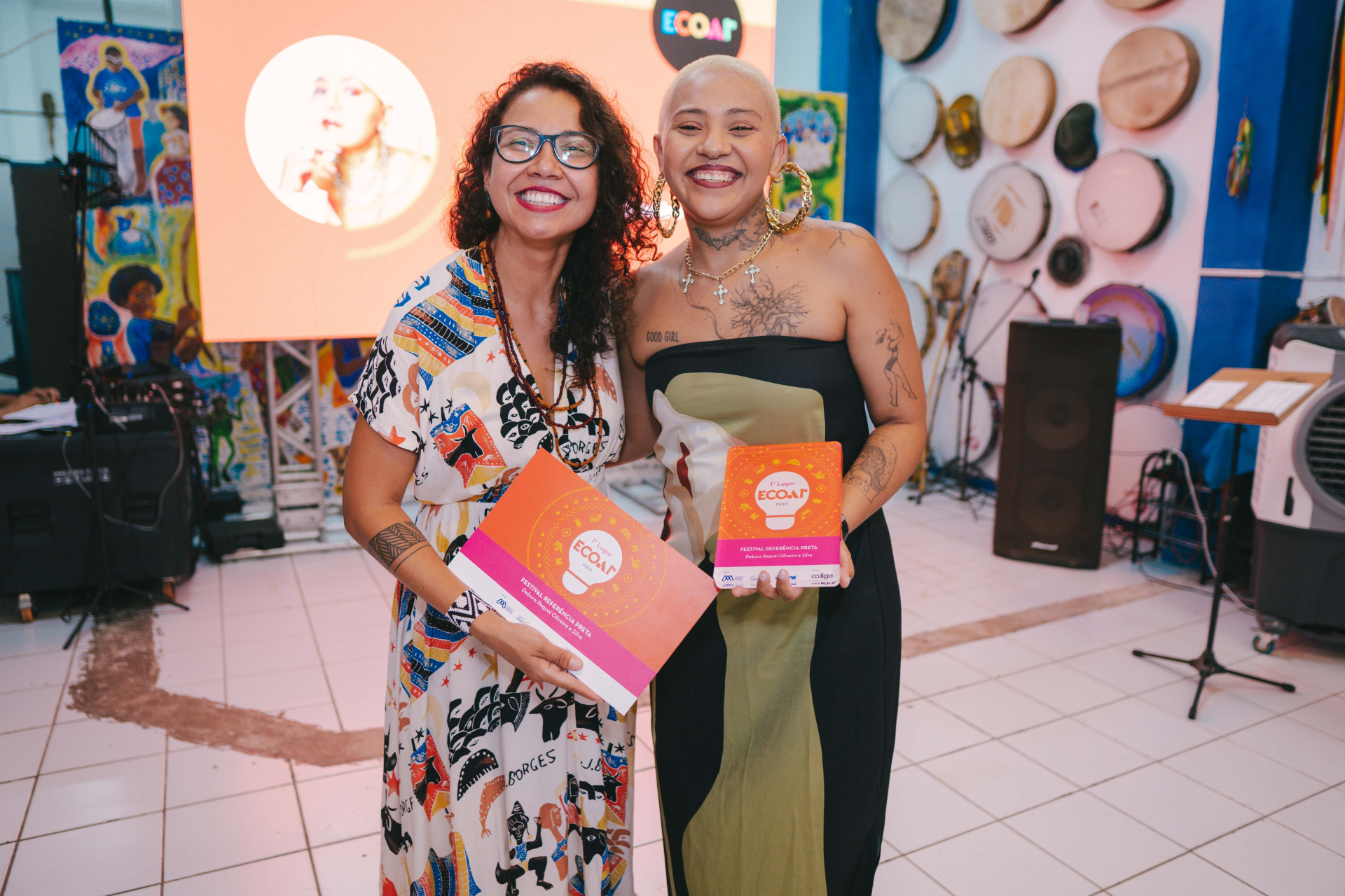 Duas mulheres posam lado a lado sorrindo para a câmera durante um evento. À esquerda, uma mulher de cabelos cacheados e óculos usa vestido estampado e segura um certificado. À direita, uma mulher de cabeça raspada, grandes brincos dourados e tatuagens aparentes usa vestido preto e verde e segura um troféu. Atrás delas, um telão com tons alaranjados exibe informações do evento “ECOAR”, enquanto instrumentos musicais decoram a parede ao fundo.