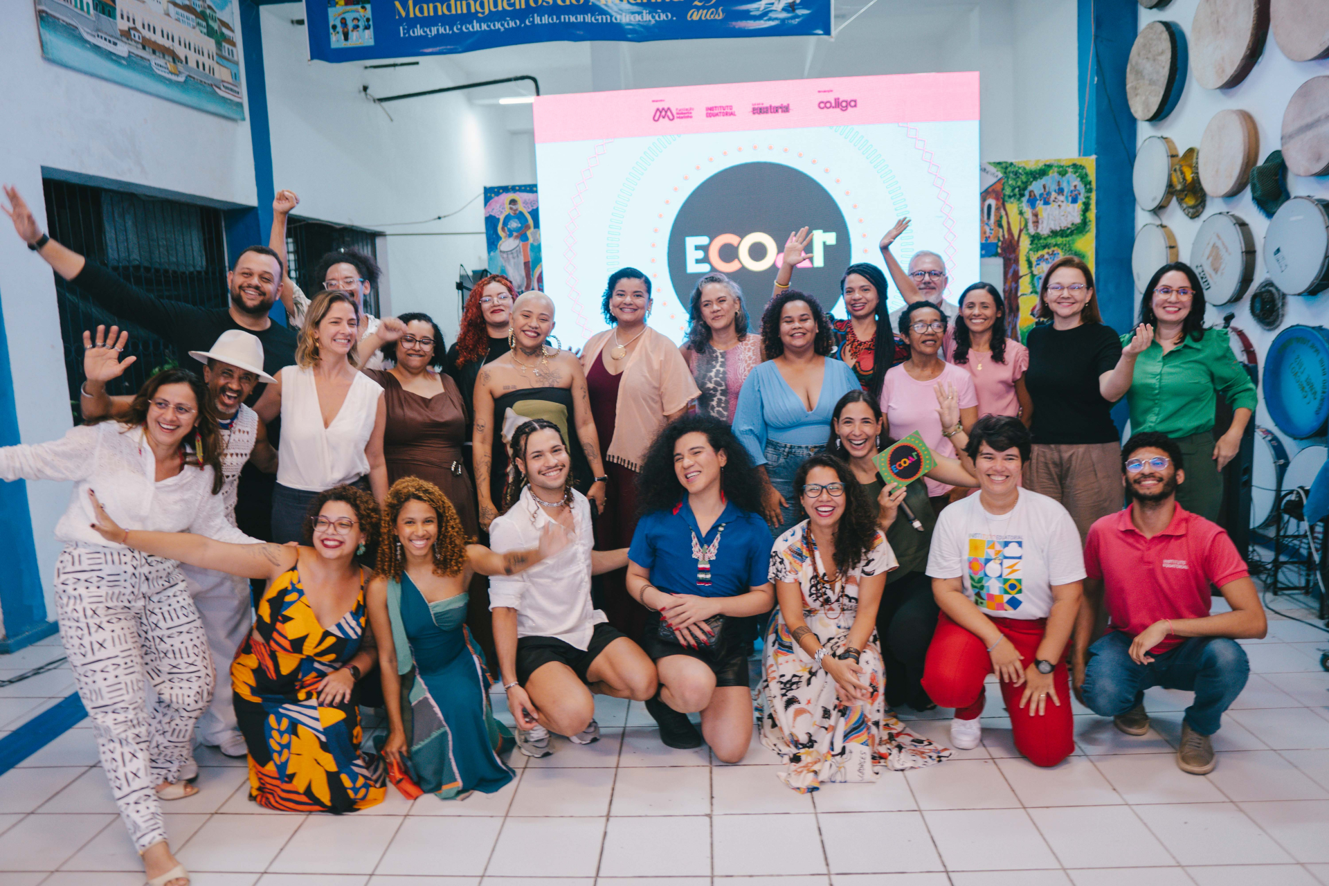 Cerca de vinte pessoas posam reunidas em um espaço cultural colorido e descontraído. O grupo é diverso em gênero, idade e aparência, com pessoas em pé e agachadas sorrindo para a câmera, algumas acenando e levantando os braços. Ao fundo, um telão iluminado exibe a palavra “ECOAR”. As paredes têm instrumentos de percussão pendurados e pinturas coloridas, criando um ambiente artístico e acolhedor.