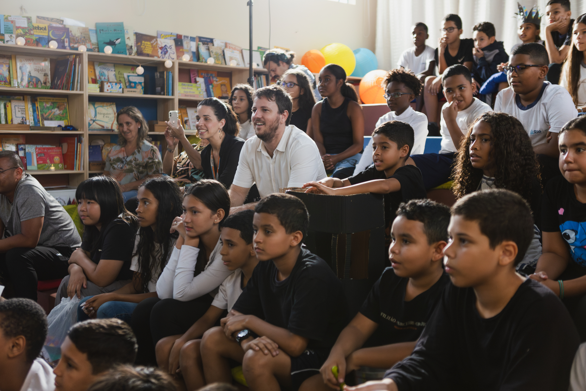 Uma sala ampla e iluminada reúne dezenas de crianças, adolescentes e alguns adultos sentados juntos em arquibancadas e almofadas, assistindo atentamente a uma atividade fora do enquadramento da foto. A maioria olha para a mesma direção com expressões de curiosidade, concentração e entusiasmo.
