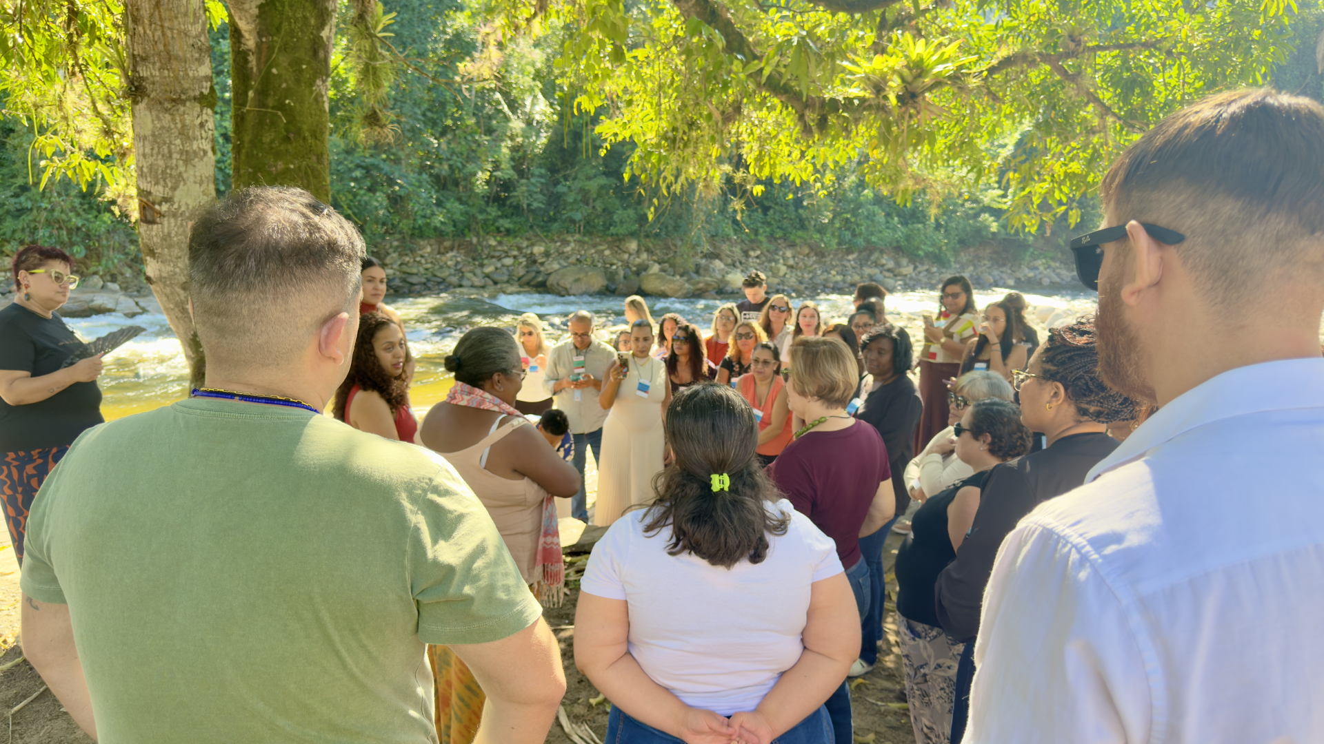 A imagem mostra o grupo ao ar livre, próximo a um rio, em um momento de roda de conversa. O cenário natural reforça a dimensão de conexão com o território e com o meio ambiente, ampliando a experiência educativa para além da sala, integrando natureza, cultura e comunidade.