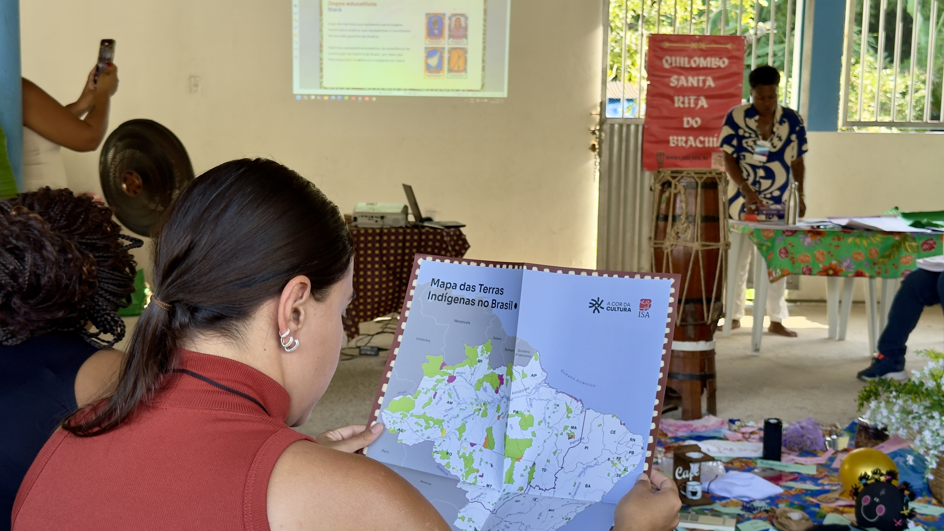 Uma participante observa um material com o título “Mapa das Terras Indígenas no Brasil”, enquanto, ao fundo, uma facilitadora conduz uma apresentação. Um banner com a inscrição “Quilombo Santa Rita do Bracuí” indica que a atividade está vinculada a um território quilombola, reforçando o contexto de educação voltada para a valorização das identidades e histórias afro-brasileiras e indígenas.