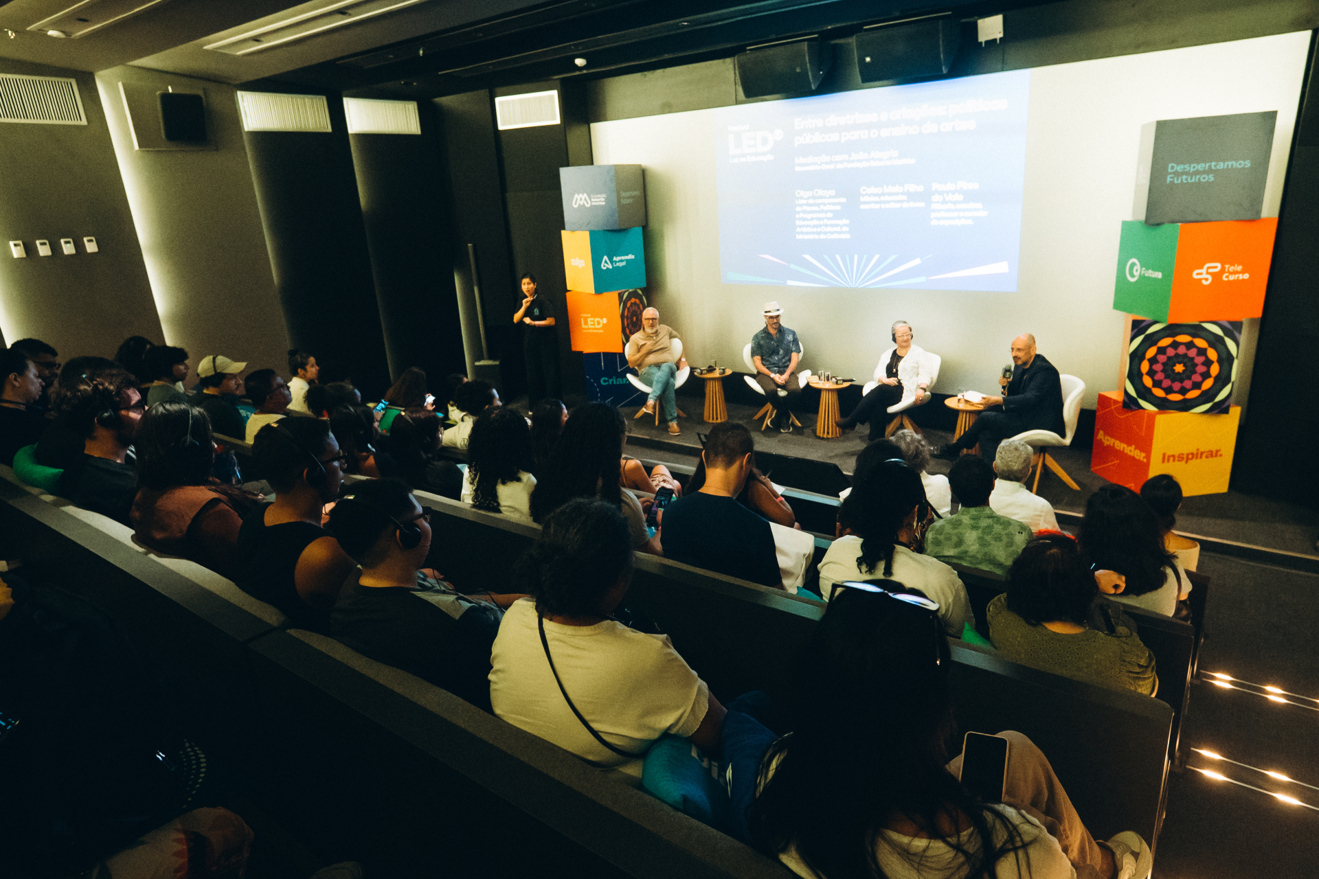 Sala de cinema ou auditório pequeno com iluminação baixa e plateia acomodada em fileiras inclinadas. No palco, quatro participantes estão sentados em poltronas brancas durante uma mesa de debate do Festival LED. Um homem fala ao microfone enquanto os demais acompanham a conversa. Atrás deles, um telão projeta o título da atividade e logos de instituições parceiras. À esquerda do palco, uma intérprete de Libras aparece em pé. O público observa atentamente, alguns usando fones de tradução simultânea.