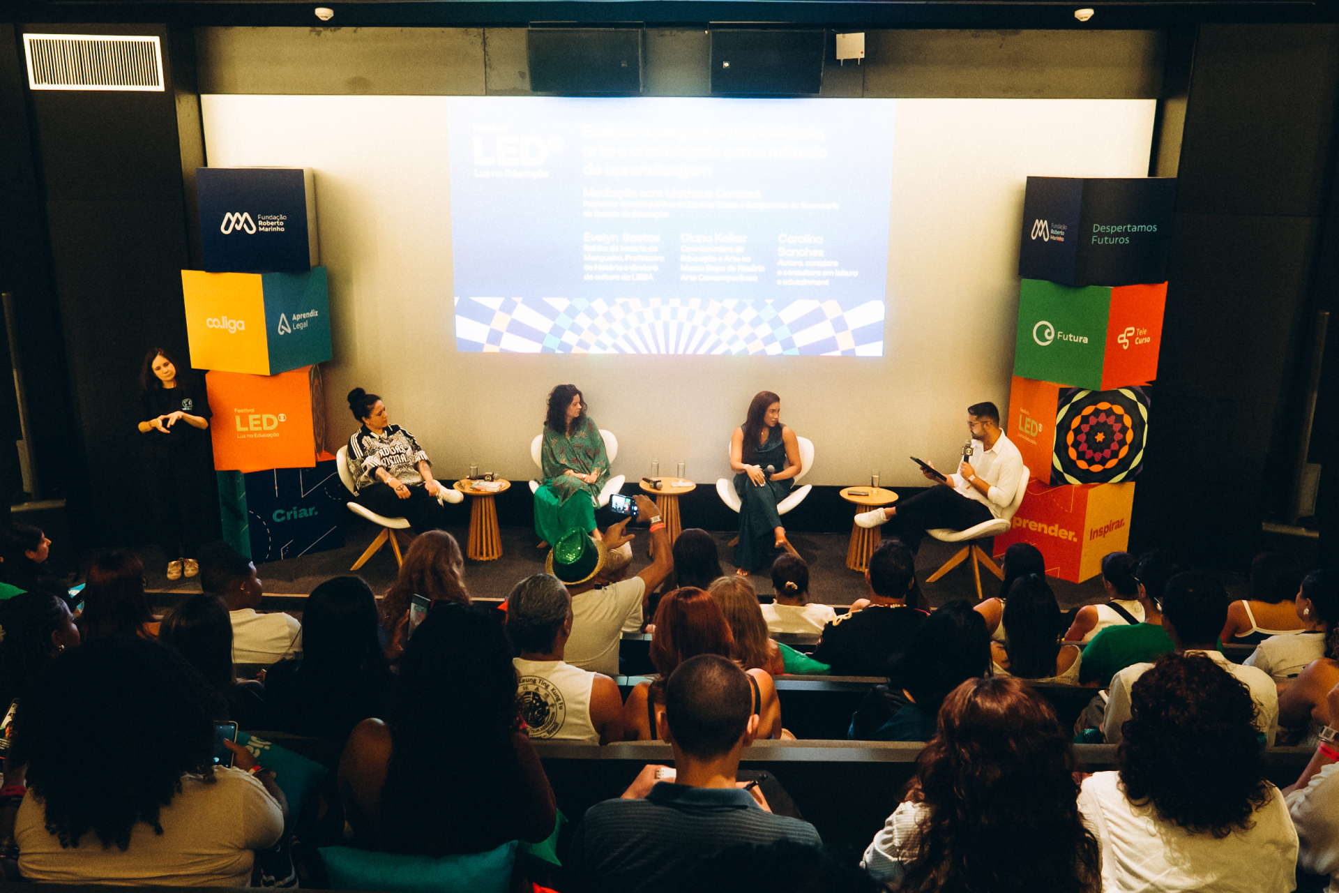 Auditório escuro e moderno durante uma mesa de debate do Festival LED – Luz na Educação. O público ocupa fileiras inclinadas voltadas para o palco, onde três mulheres e um mediador estão sentados em cadeiras brancas, conversando diante da plateia. O homem à direita segura um microfone e um tablet enquanto conduz a conversa.