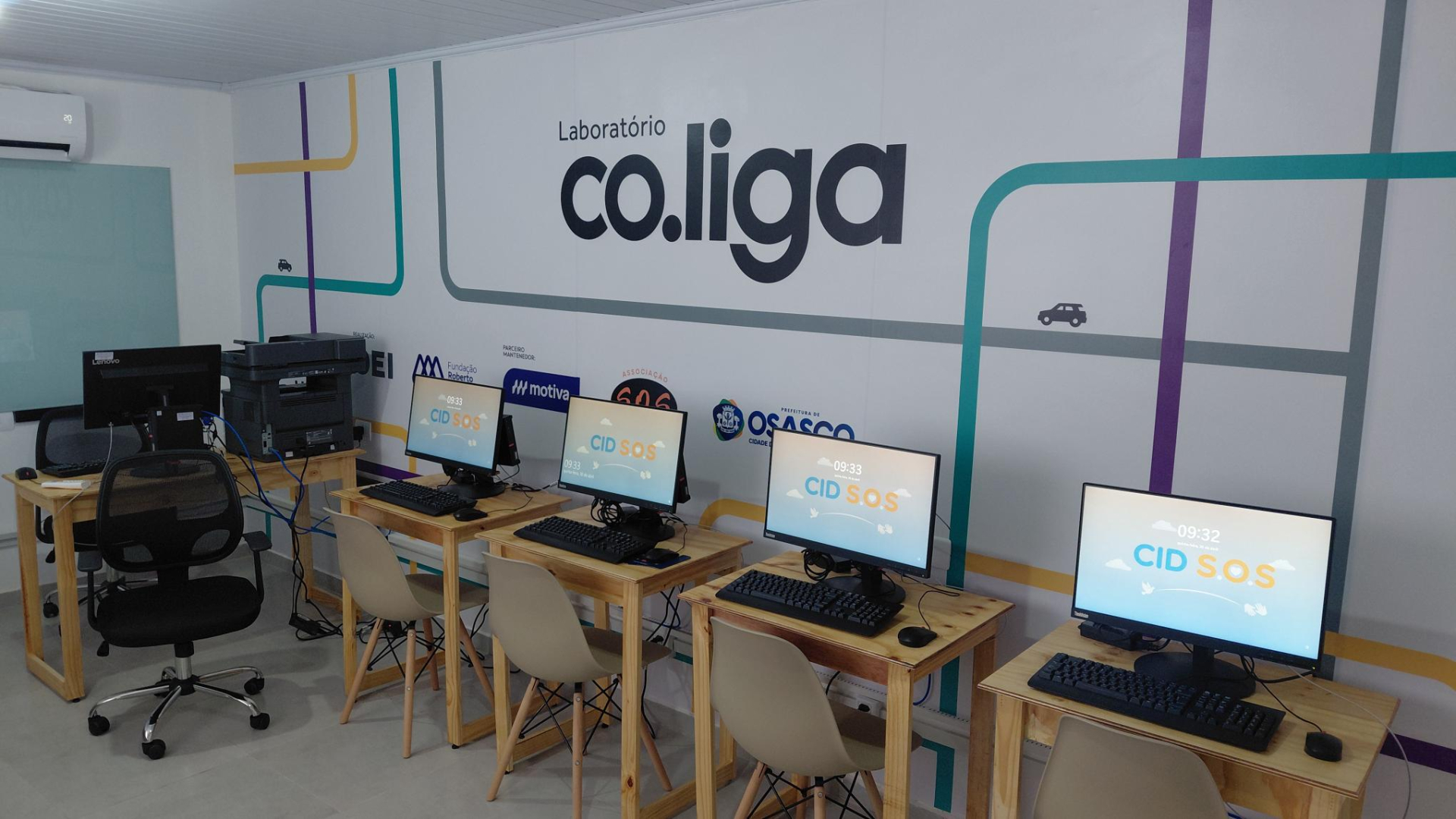 A imagem mostra um laboratório de informática organizado e moderno, identificado como “Laboratório co.liga” em destaque na parede ao fundo. O espaço conta com várias estações de trabalho alinhadas, cada uma equipada com computador, teclado e mouse, dispostas sobre mesas de madeira clara com cadeiras simples.