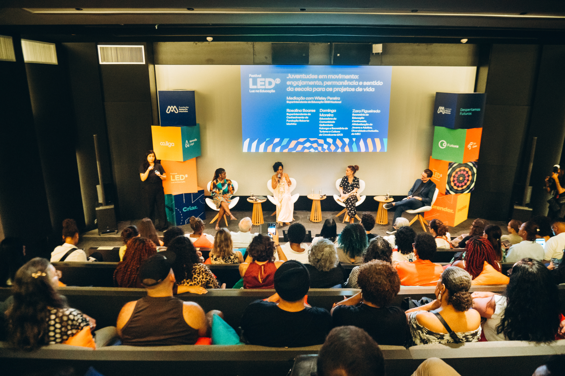 Auditório moderno com iluminação baixa e plateia distribuída em fileiras inclinadas, voltadas para um palco onde acontece uma mesa de debate do Festival LED – Luz na Educação. No centro do palco, quatro participantes estão sentados em cadeiras brancas, conversando diante do público. Uma das mulheres fala ao microfone enquanto os demais acompanham atentos.