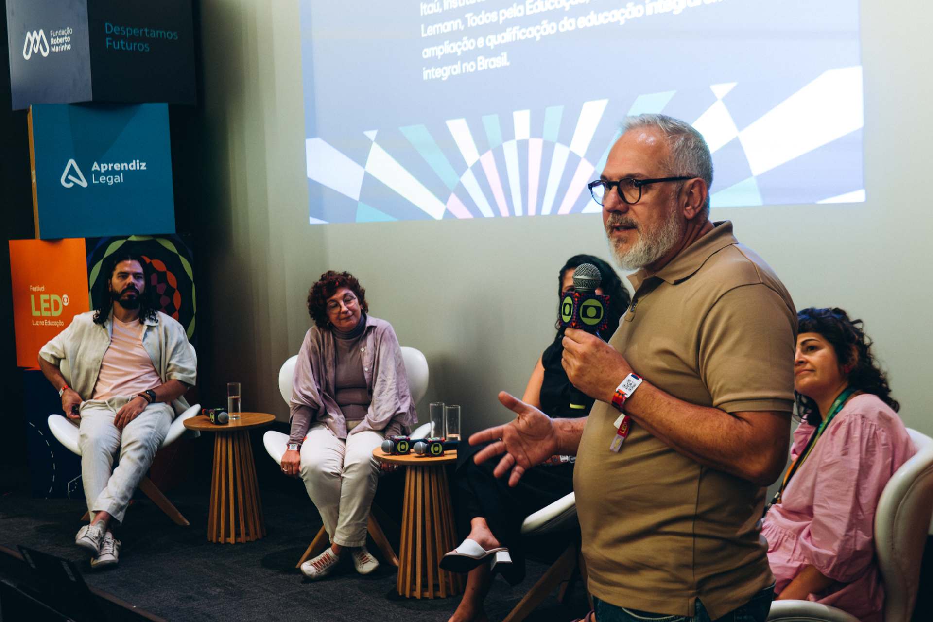 Fotografia de um debate em auditório durante o Festival LED. Em primeiro plano, um homem branco de cabelos grisalhos curtos, barba branca e óculos fala ao microfone enquanto está de pé no palco. Ele veste camisa polo bege e gesticula com uma das mãos, em uma postura de fala e interação com o público.  Ao fundo, outras quatro pessoas participam da mesa de conversa, sentadas em poltronas brancas ao redor de pequenas mesas de madeira. 