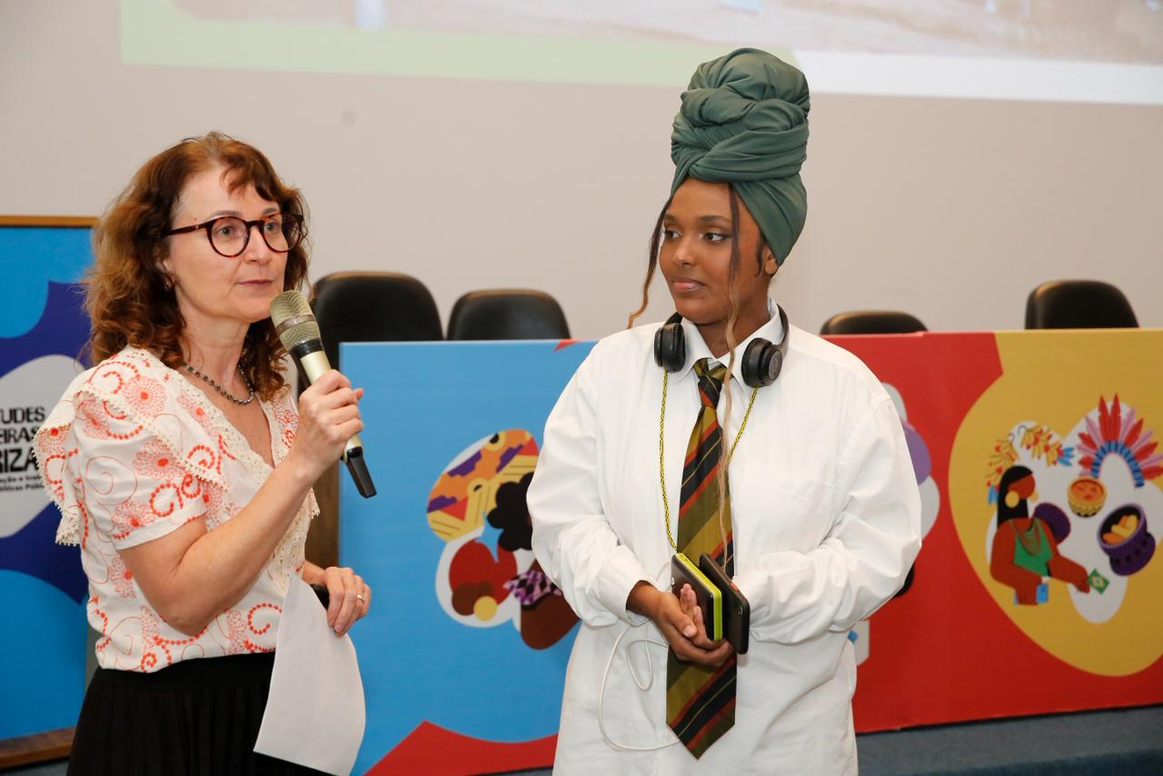 Fotografia de um evento em ambiente interno. À esquerda, uma mulher branca de meia-idade, com cabelos castanhos ondulados e óculos, fala ao microfone enquanto segura algumas folhas de papel. Ela veste uma blusa clara com detalhes bordados em vermelho e uma saia preta.  Ao lado dela, uma jovem negra observa atentamente a fala. Ela usa um turbante verde grande enrolado na cabeça, camisa branca oversized e gravata listrada em tons escuros.
