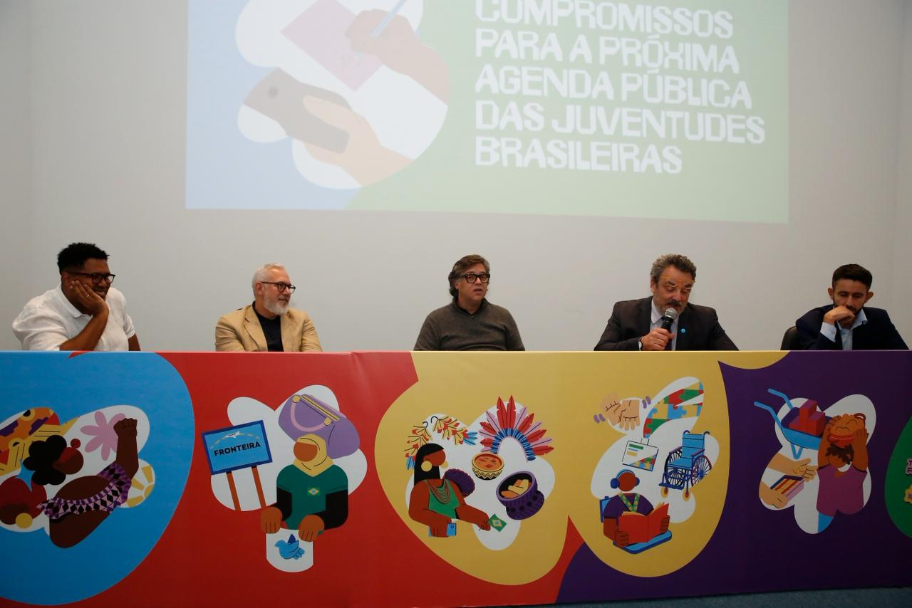 Em um auditório, cinco homens participam de uma mesa de debate sobre juventudes brasileiras. Eles estão sentados atrás de uma bancada longa e colorida, ilustrada com desenhos que representam diversidade, inclusão e participação social. Entre as imagens aparecem pessoas indígenas, símbolos de acessibilidade, leitura, tecnologia e manifestações culturais.
