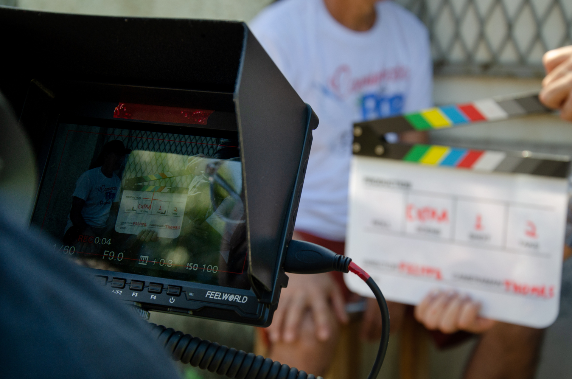 A imagem mostra um momento de produção audiovisual em andamento. Em primeiro plano, um monitor de câmera exibe a gravação de uma cena, enquanto, ao fundo, uma claquete cinematográfica é posicionada diante dos participantes para marcar o início da tomada. As mãos e partes do corpo das pessoas envolvidas aparecem parcialmente desfocadas, direcionando a atenção para os equipamentos de filmagem e para o processo técnico da gravação.