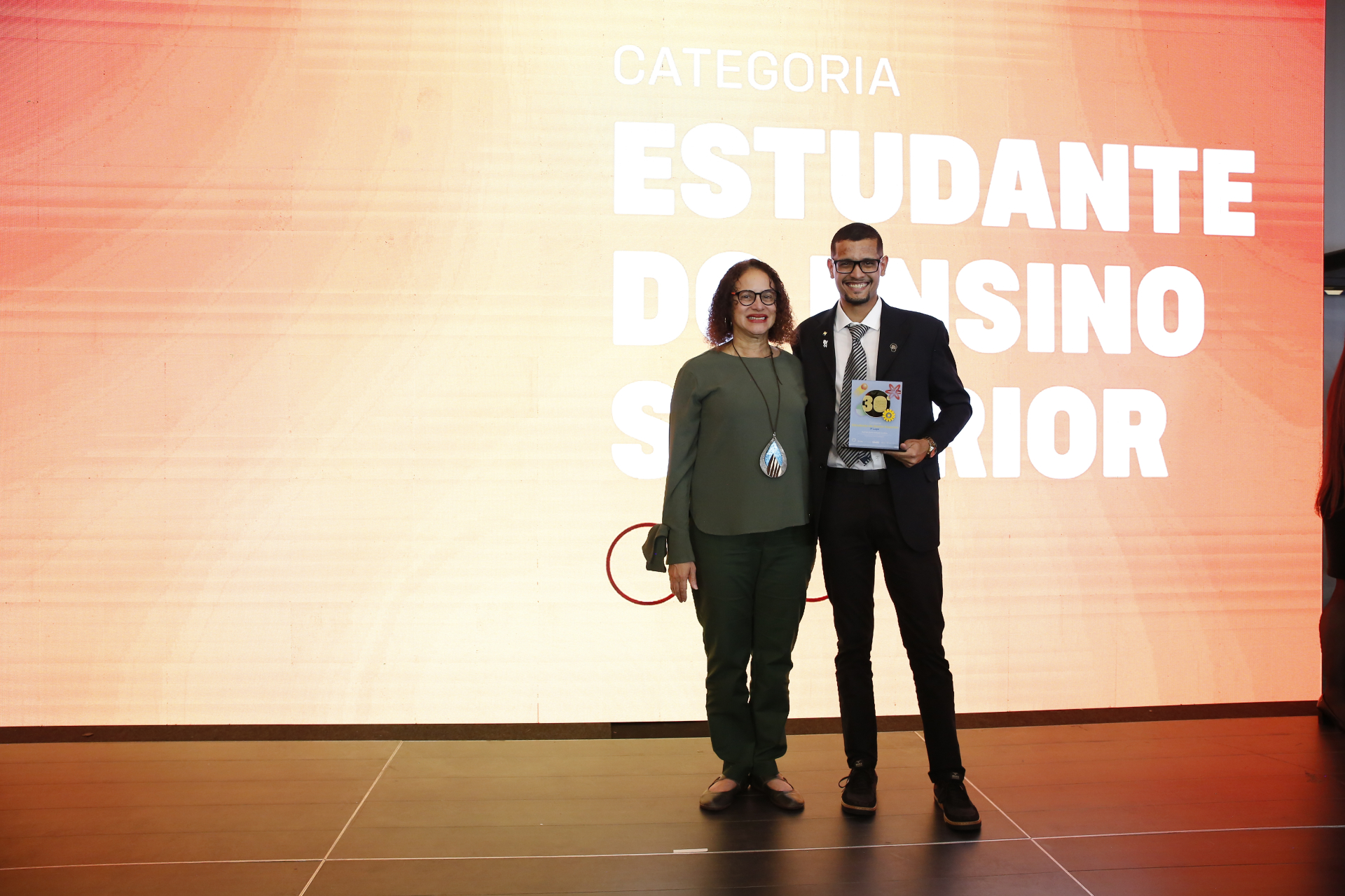 Em um palco de premiação, duas pessoas posam lado a lado para uma fotografia. À esquerda, uma mulher usa blusa verde de mangas compridas, calça escura e óculos. À direita, um jovem veste terno escuro, camisa branca e gravata listrada, sorrindo enquanto segura um troféu ou placa de homenagem.