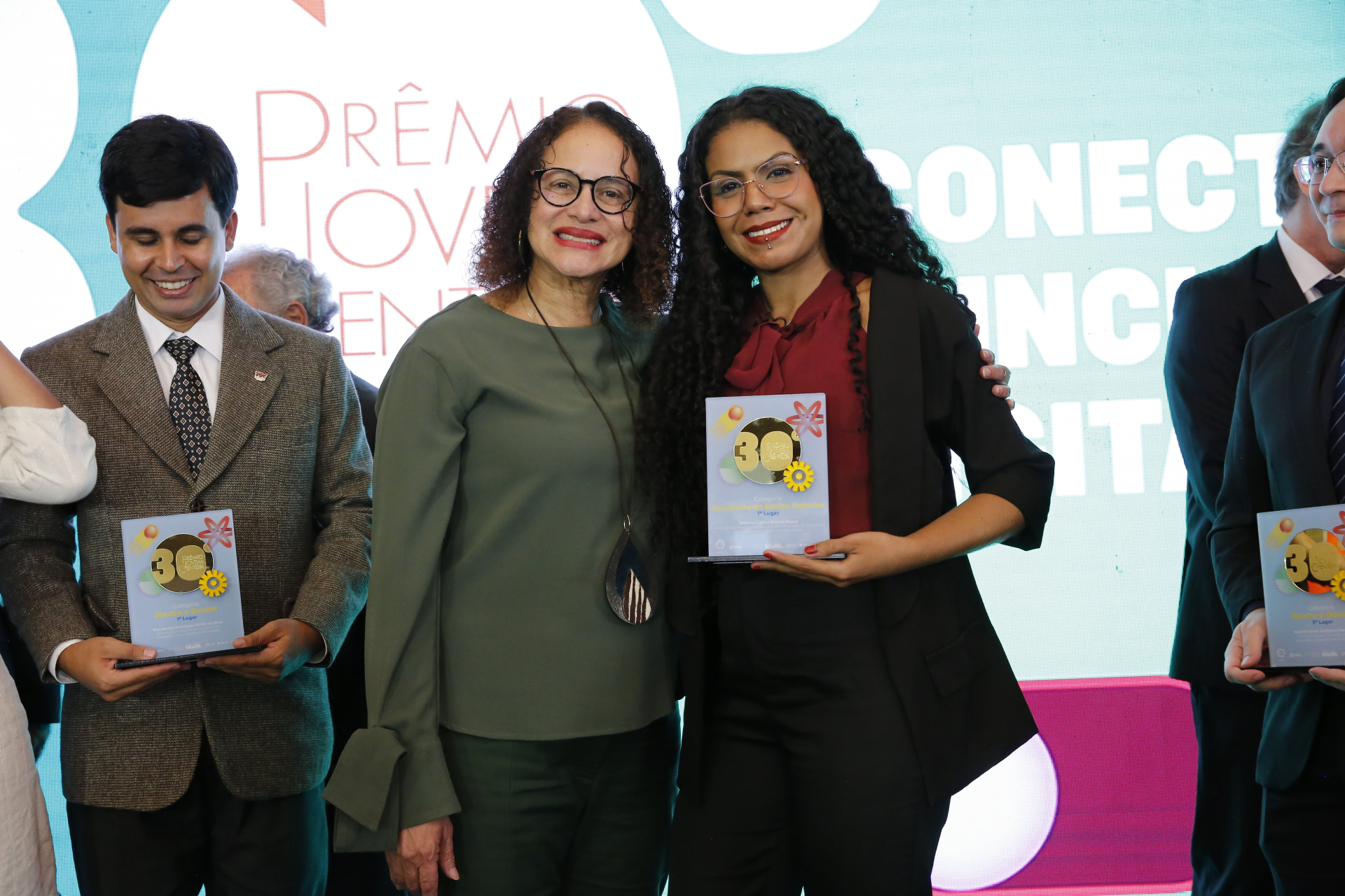 Fotografia registrada durante uma cerimônia de premiação. No centro da imagem, duas mulheres posam sorrindo para a câmera. À esquerda, uma mulher de cabelos cacheados curtos, óculos e roupa verde escura mantém um dos braços ao redor da premiada. À direita, uma jovem de cabelos longos e cacheados, usando óculos, blusa vinho com laço na gola e blazer preto, segura uma placa de reconhecimento.