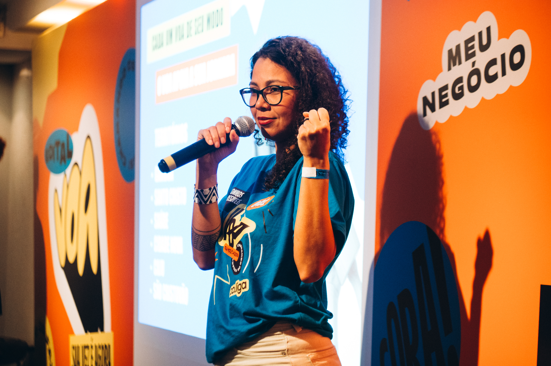 Retrato de uma mulher de cabelos cacheados e óculos falando ao microfone durante uma apresentação. Ela veste camiseta azul do evento e gesticula com uma das mãos enquanto se dirige ao público. Ao fundo, aparecem uma tela de projeção e painéis laranja com palavras relacionadas ao empreendedorismo e à formação. A iluminação destaca a palestrante, transmitindo confiança, protagonismo e engajamento.