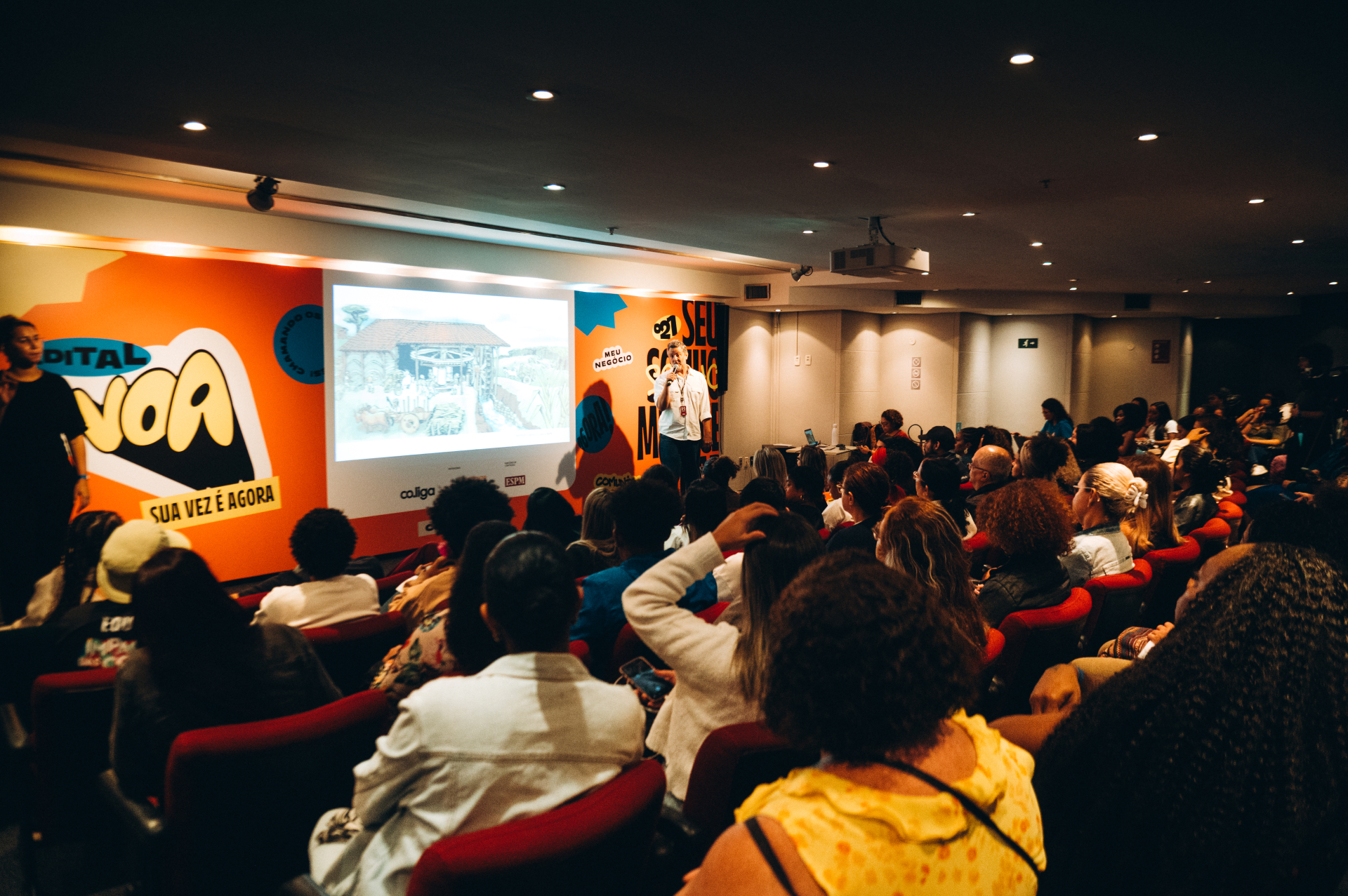 Vista ampla de um auditório lotado durante uma apresentação. O público ocupa quase todas as cadeiras vermelhas e está voltado para o palco. Um palestrante fala ao microfone ao lado de uma projeção exibida em tela grande. A parede do palco é decorada com elementos gráficos coloridos e frases do evento. A imagem destaca a grande participação do público e o ambiente de troca de conhecimentos.
