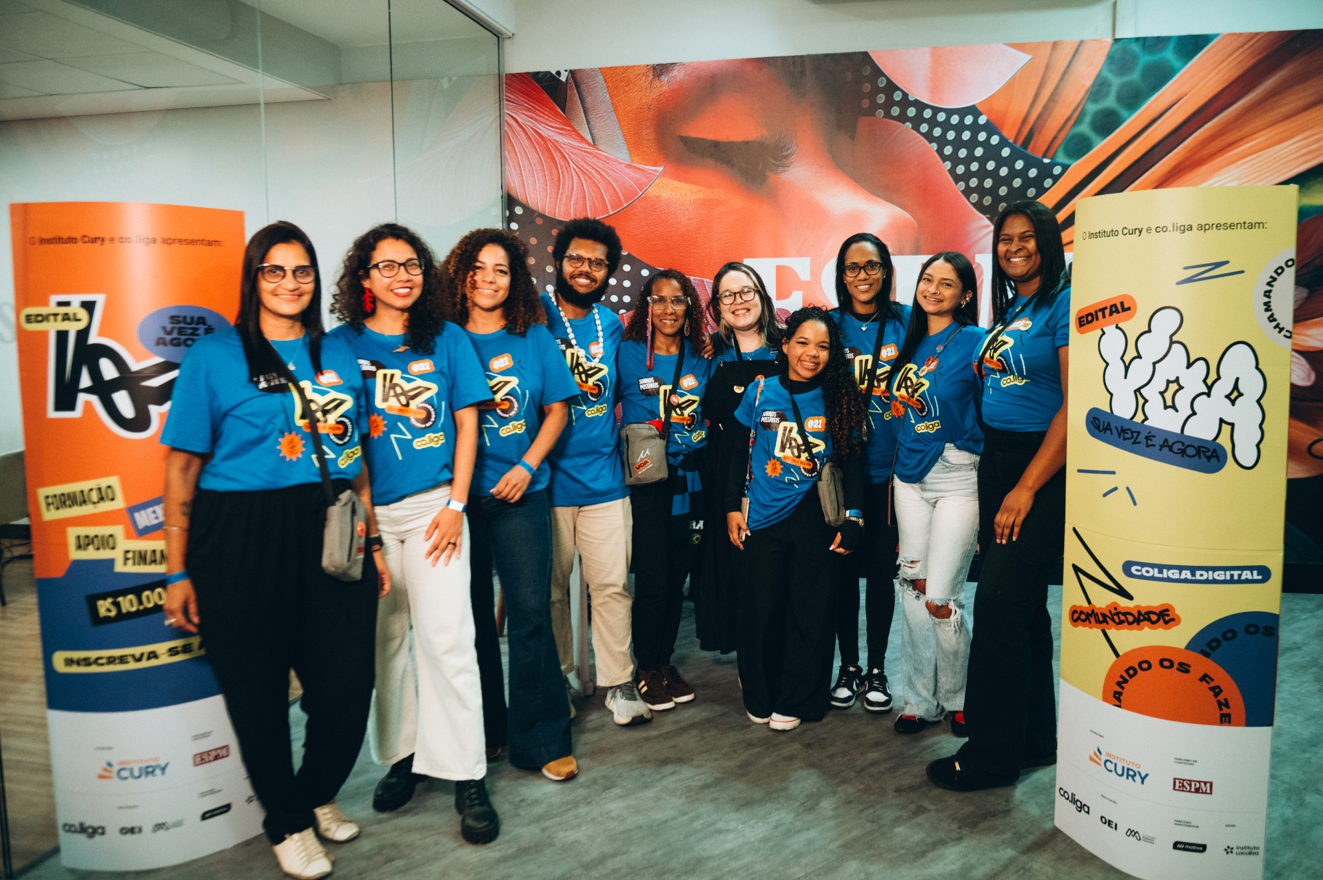 Fotografia de um grupo de dez pessoas posando para uma foto em um espaço interno de eventos. A maioria veste camisetas azuis com a identidade visual do programa. Todos estão sorrindo e posicionados entre dois totens coloridos do edital, com predominância das cores laranja, azul e amarelo. Ao fundo, há um grande painel artístico com ilustrações e grafismos em tons quentes.
