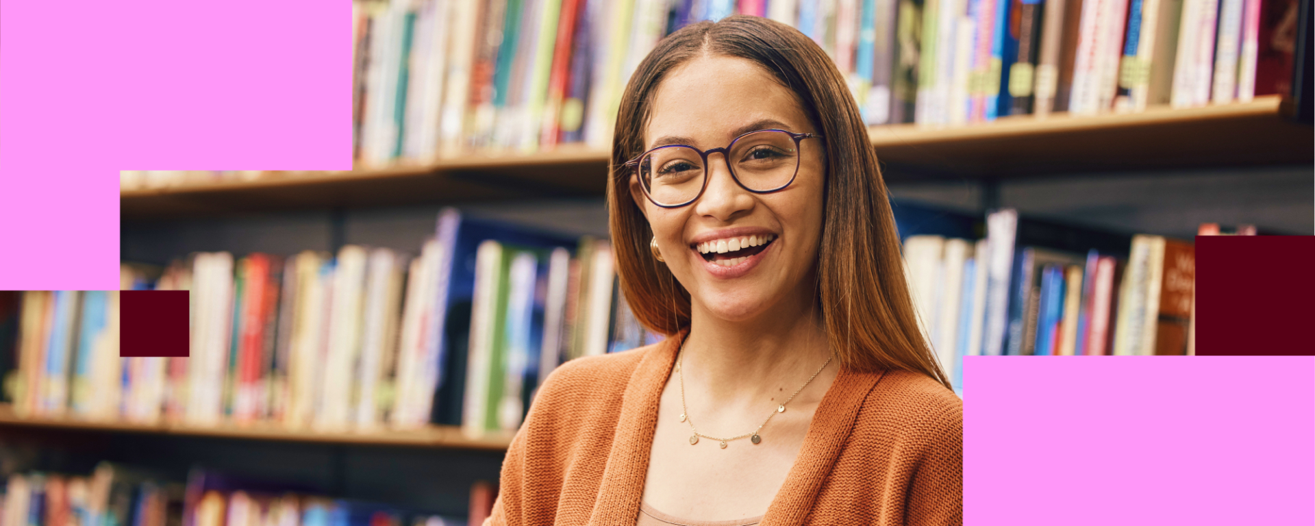 Fotografia em formato horizontal mostrando uma jovem sorrindo em uma biblioteca. Ela está posicionada à direita da imagem, olhando diretamente para a câmera. Tem cabelos longos e lisos, castanho-escuros, usa óculos de armação arredondada e veste uma blusa em tom terracota. Também usa um colar delicado dourado.  Ao fundo, aparecem estantes repletas de livros, desfocadas, criando um ambiente acolhedor e associado ao estudo e à educação. A iluminação é suave e quente, destacando a expressão alegre e receptiva.