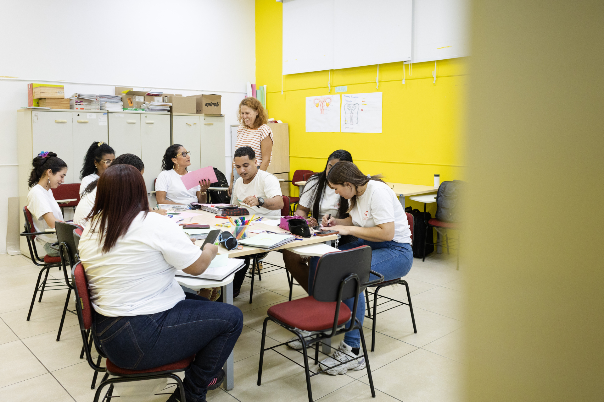 Fotografia de uma sala de aula durante uma atividade em grupo. Oito estudantes estão sentados ao redor de uma mesa retangular, trabalhando em conjunto com cadernos, folhas de papel coloridas, canetas, lápis e outros materiais de escrita espalhados sobre a mesa. A maioria veste camisetas brancas e está concentrada em escrever, desenhar ou conversar sobre a atividade.