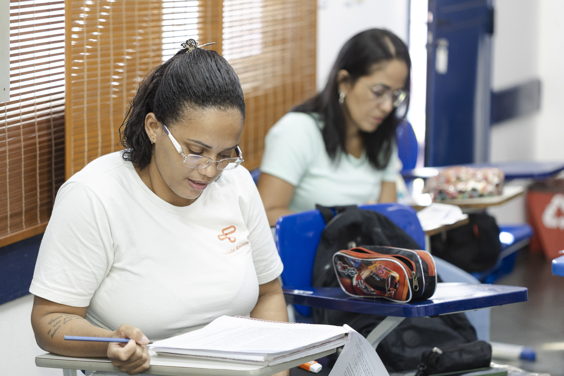 Em primeiro plano, uma mulher de cabelos presos e óculos segura um lápis enquanto lê atentamente um caderno aberto sobre a carteira. Ela veste uma camiseta branca e demonstra expressão de concentração.  Ao fundo, outra estudante, também usando óculos, está sentada em uma carteira individual, com o olhar voltado para seu material de estudo. Sobre a mesa há um estojo colorido, cadernos e outros materiais escolares.