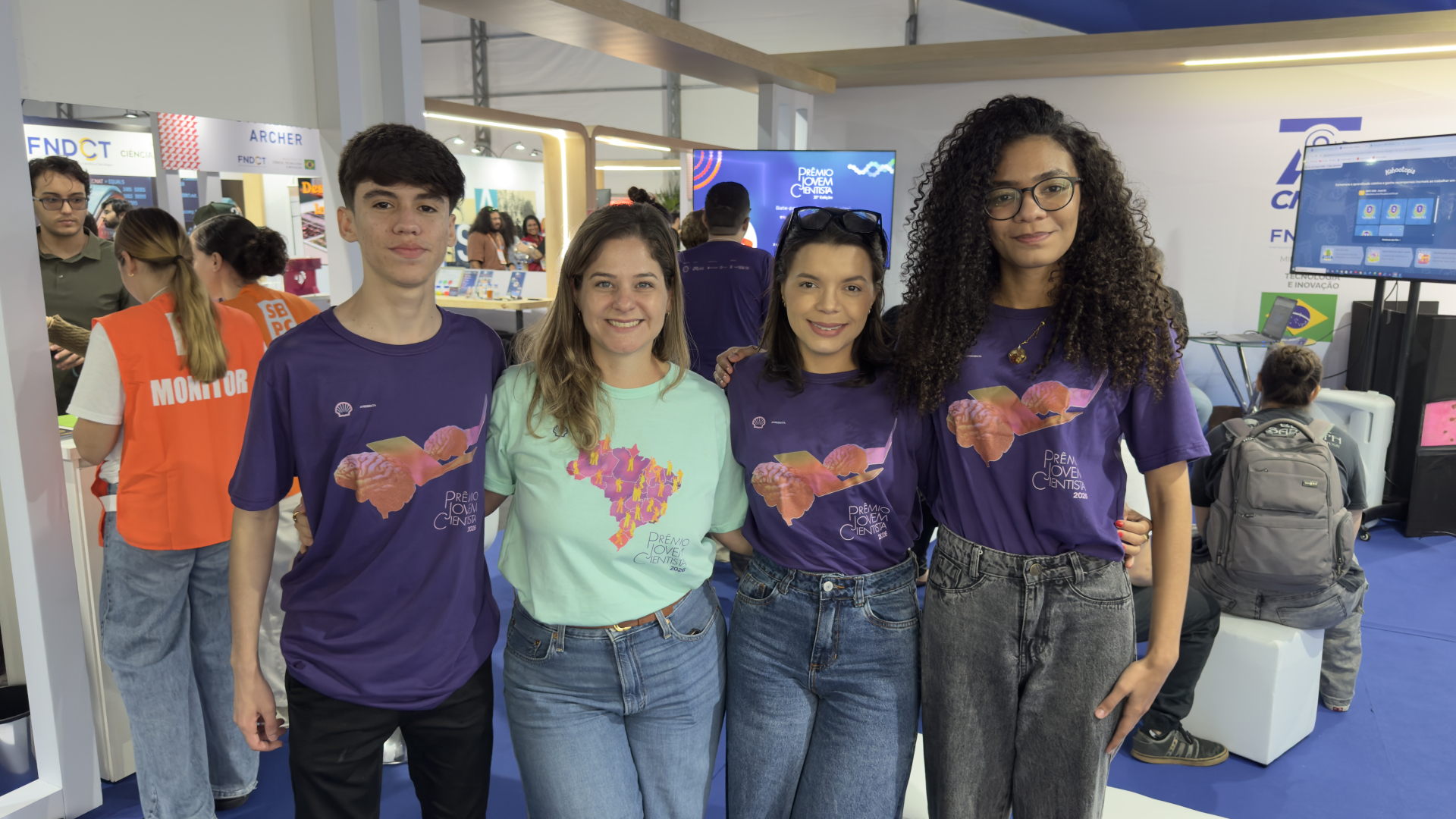 Fotografia de quatro pessoas posando lado a lado em um estande de evento voltado para ciência e inovação. O grupo sorri para a câmera, com os braços apoiados nos ombros ou nas costas uns dos outros, transmitindo um clima de integração.  Três participantes vestem camisetas roxas do Prêmio Jovem Cientista, com uma ilustração de um cérebro e um livro. Ao centro, uma mulher usa uma camiseta verde-clara com a identidade visual do prêmio e uma estampa do mapa do Brasil.