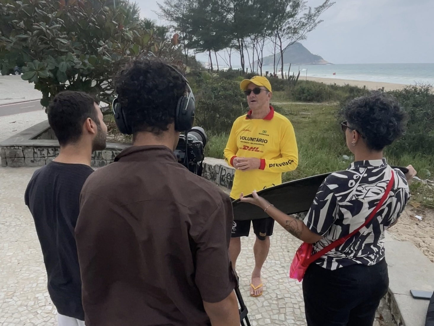 Uma equipe de produção audiovisual grava uma entrevista ao ar livre, em um calçadão à beira-mar. Em primeiro plano, três pessoas operam os equipamentos de filmagem: uma utiliza uma câmera profissional, outra monitora a captação de áudio com fones de ouvido e uma terceira segura um rebatedor de luz para melhorar a iluminação da cena. À frente da equipe, uma pessoa vestindo camiseta amarela de voluntário concede a entrevista, de pé e voltada para a câmera.
