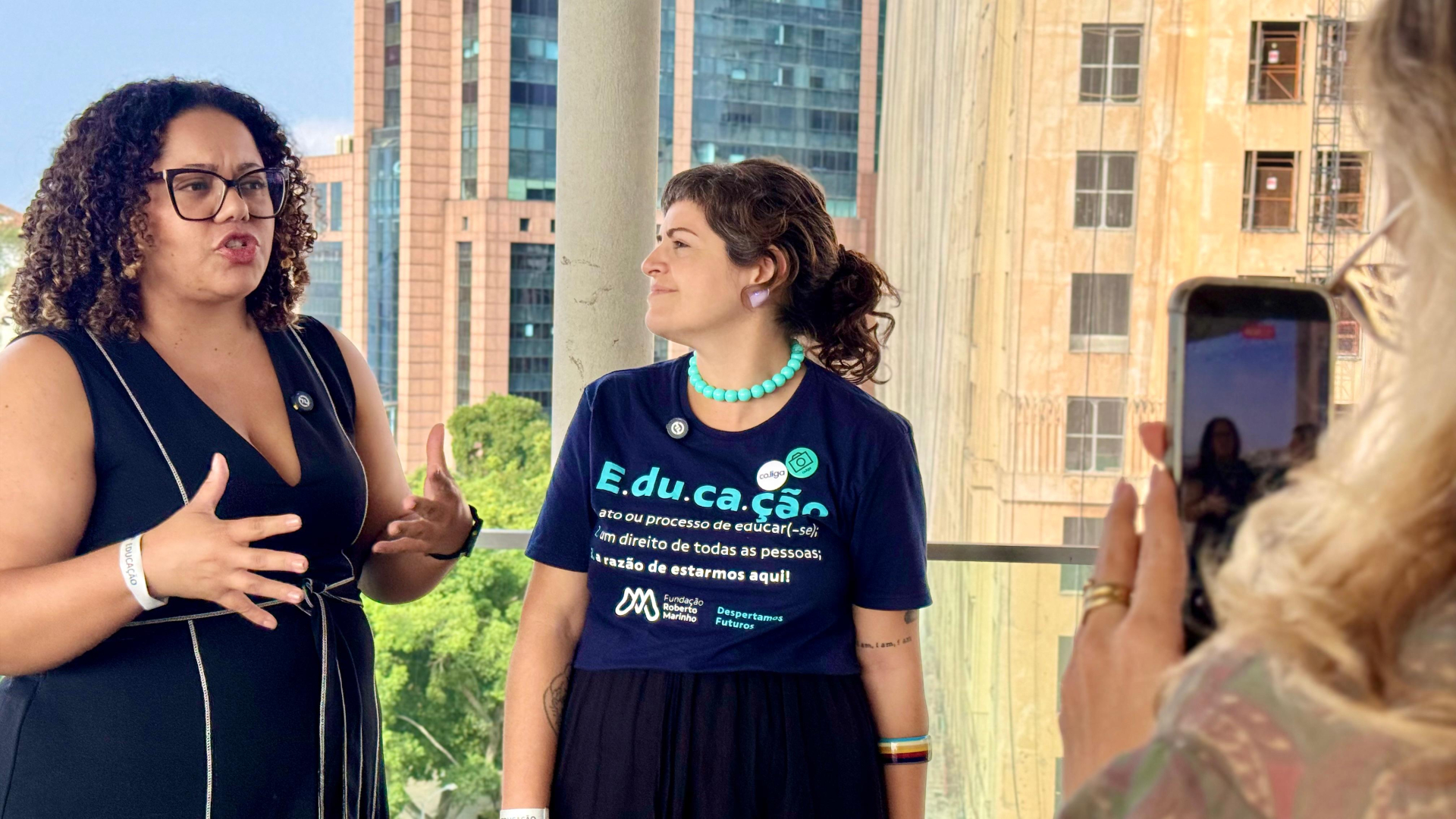 Fotografia de uma entrevista sendo gravada em um espaço aberto com vista para prédios ao fundo. À esquerda, uma mulher de cabelos cacheados, óculos e vestido azul-marinho fala enquanto gesticula com as mãos, demonstrando expressão atenta e envolvida. Ao seu lado, outra mulher sorri e acompanha a conversa. Ela veste uma camiseta azul-marinho com a palavra "Educação" em destaque e a identidade visual da Fundação Roberto Marinho, além de colar e brincos coloridos.