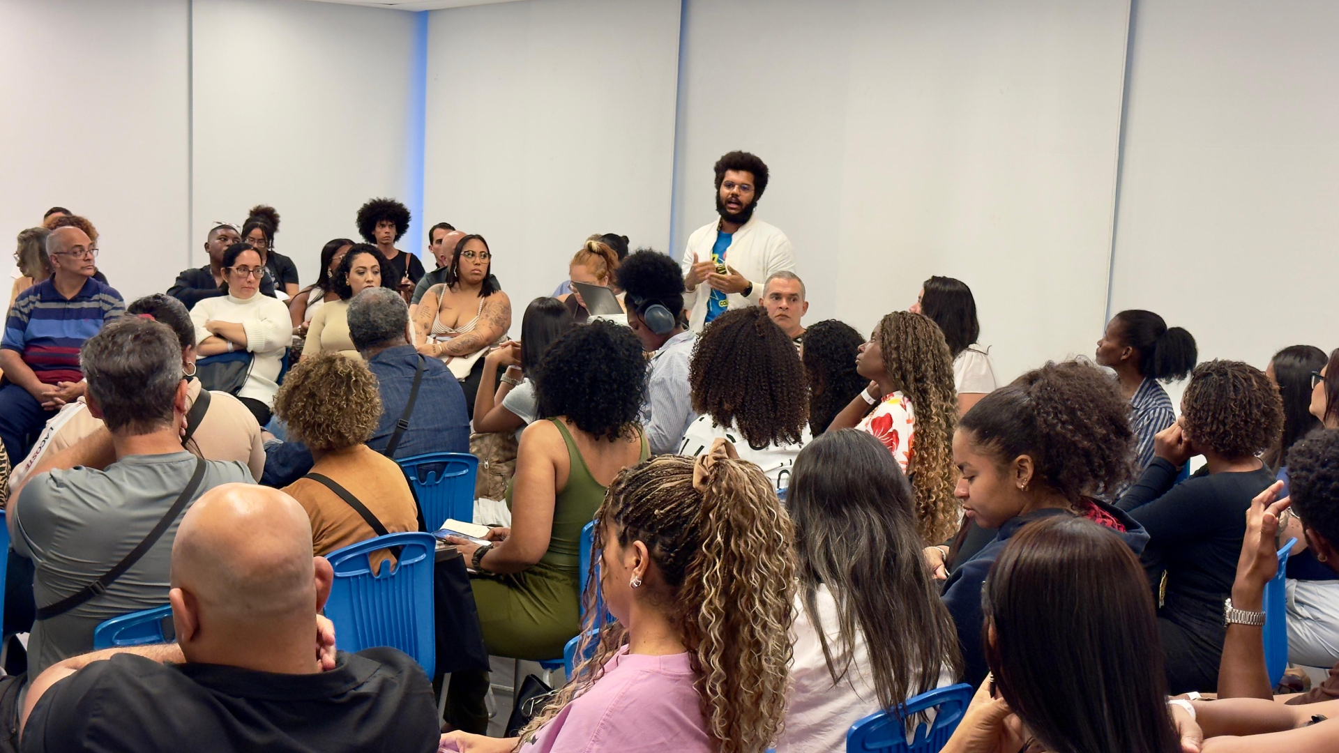 A imagem mostra uma atividade em formato de roda de conversa ou plenária, realizada em uma sala de eventos ou treinamento.  No centro da cena, um homem em pé faz uma intervenção, aparentemente compartilhando uma pergunta, comentário ou relato com os demais participantes. Ele gesticula com as mãos enquanto fala, e todos ao seu redor direcionam a atenção para ele. Ao fundo, outras pessoas acompanham a fala de forma atenta.  O público é numeroso e diverso, reunindo homens e mulheres de diferentes idades.