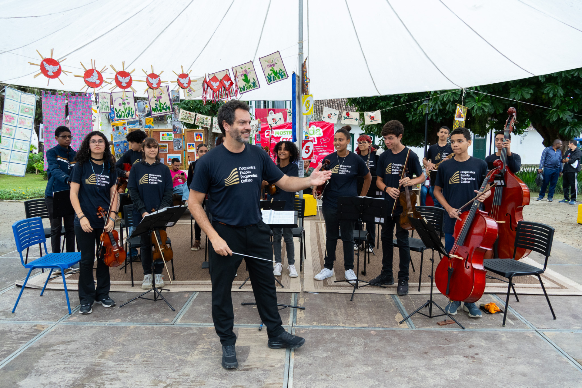 Fotografia de uma apresentação musical ao ar livre. Em primeiro plano, um maestro está no centro da cena, sorrindo e gesticulando com uma batuta enquanto conduz uma orquestra formada por crianças e adolescentes. Os integrantes usam camisetas pretas com a identificação da Orquestra Escola Pequena Calixto e seguram instrumentos de cordas, como violinos, violas e contrabaixos