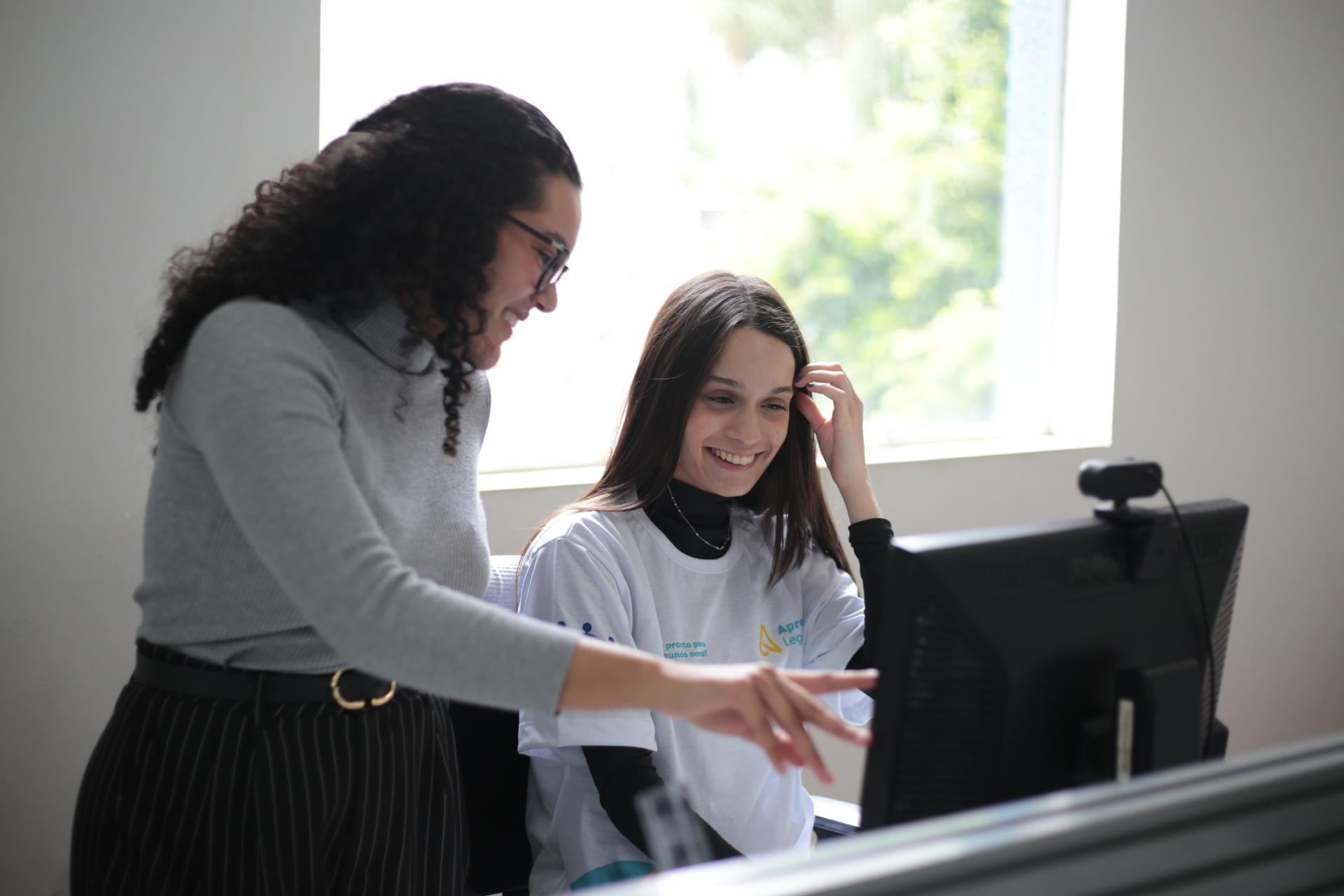 Educadora acompanha uma jovem durante uma atividade em frente ao computador. As duas observam a tela e conversam sobre a tarefa realizada.