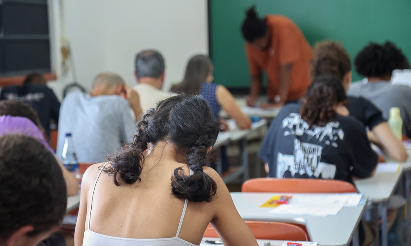 A imagem mostra uma sala de aula com estudantes sentados em suas cadeiras, de costas e olhando para a mesa. Uma professora aparece o fundo na frente de um quadro negro. 