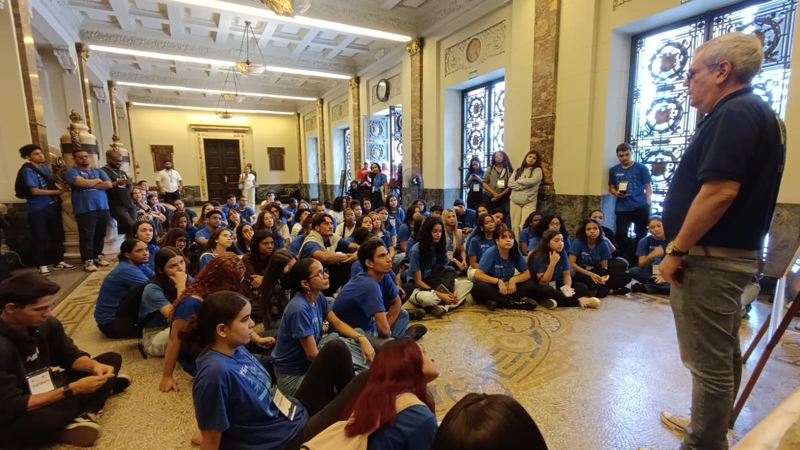 Um grande grupo de jovens com camisas azuis está sentado no chão com piso de mosaico de mármore em um grande e imponente salão. Eles estão voltados para a direita, ouvindo um homem mais velho, de pé, também com uma camisa azul. O salão tem piso de mosaico ornamentado, colunas altas de mármore e portas e janelas de madeira escura ao fundo. A iluminação é natural e quente.