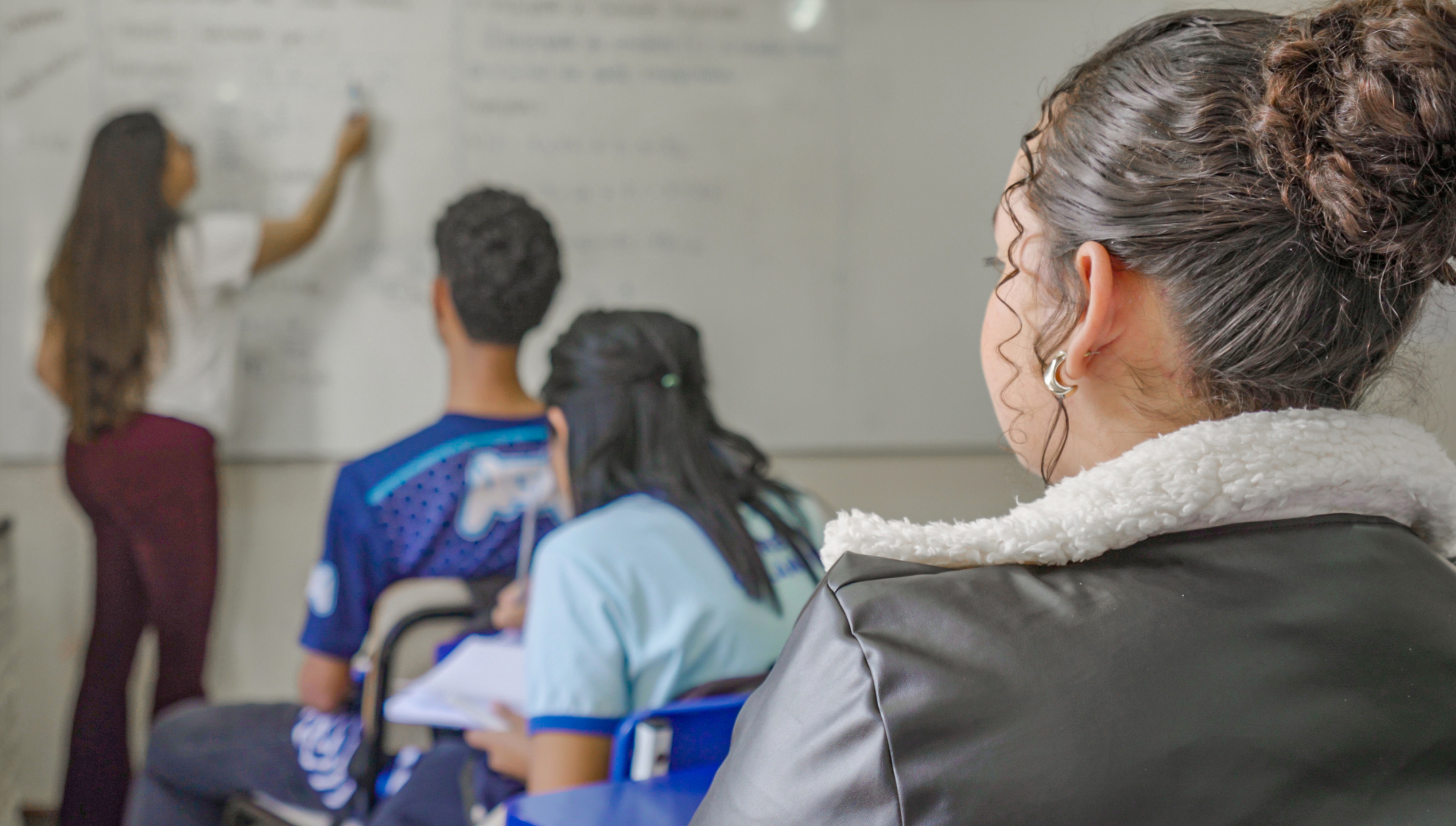 mostra uma atividade em sala de aula. Em primeiro plano, uma estudante acompanha atentamente o que acontece à frente, enquanto outros alunos estão sentados e voltados para um quadro branco. Ao fundo, uma estudante escreve no quadro, aparentemente conduzindo ou apresentando uma atividade para a turma. Alguns alunos seguram cadernos ou folhas, indicando um momento de estudo, discussão ou trabalho coletivo.