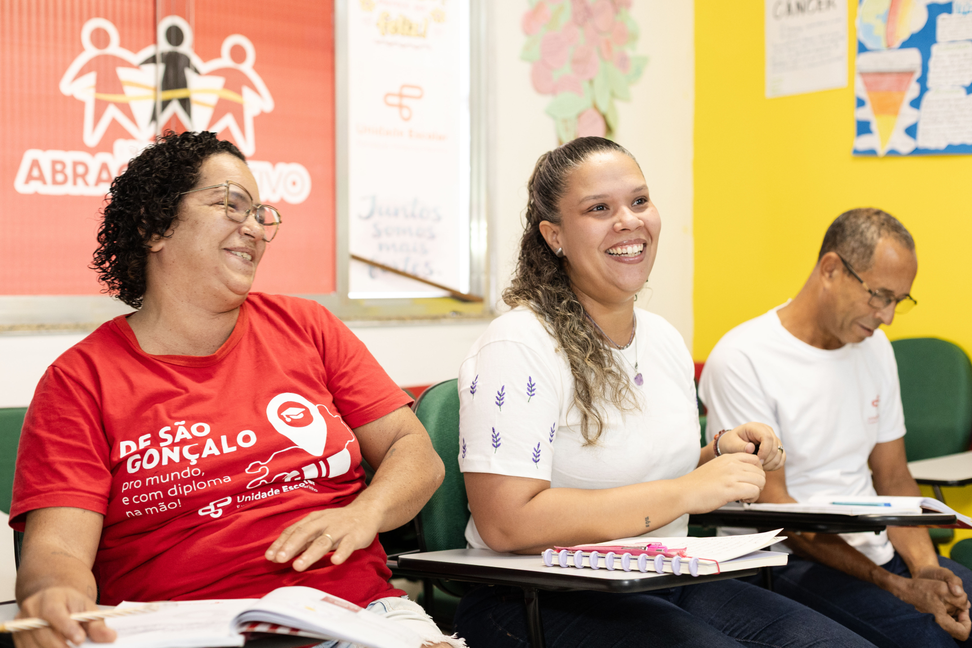 Fotografia em plano médio de três adultos sentados lado a lado em carteiras escolares. À esquerda, uma mulher sorri de camiseta vermelha. Ao centro, outra mulher sorri de camiseta branca, cabelos presos e caderno aberto. À direita, um homem de óculos e camiseta branca olha para baixo, atento. Ao fundo, uma parede amarela e vermelha com cartazes educativos. Iluminação clara e focada.