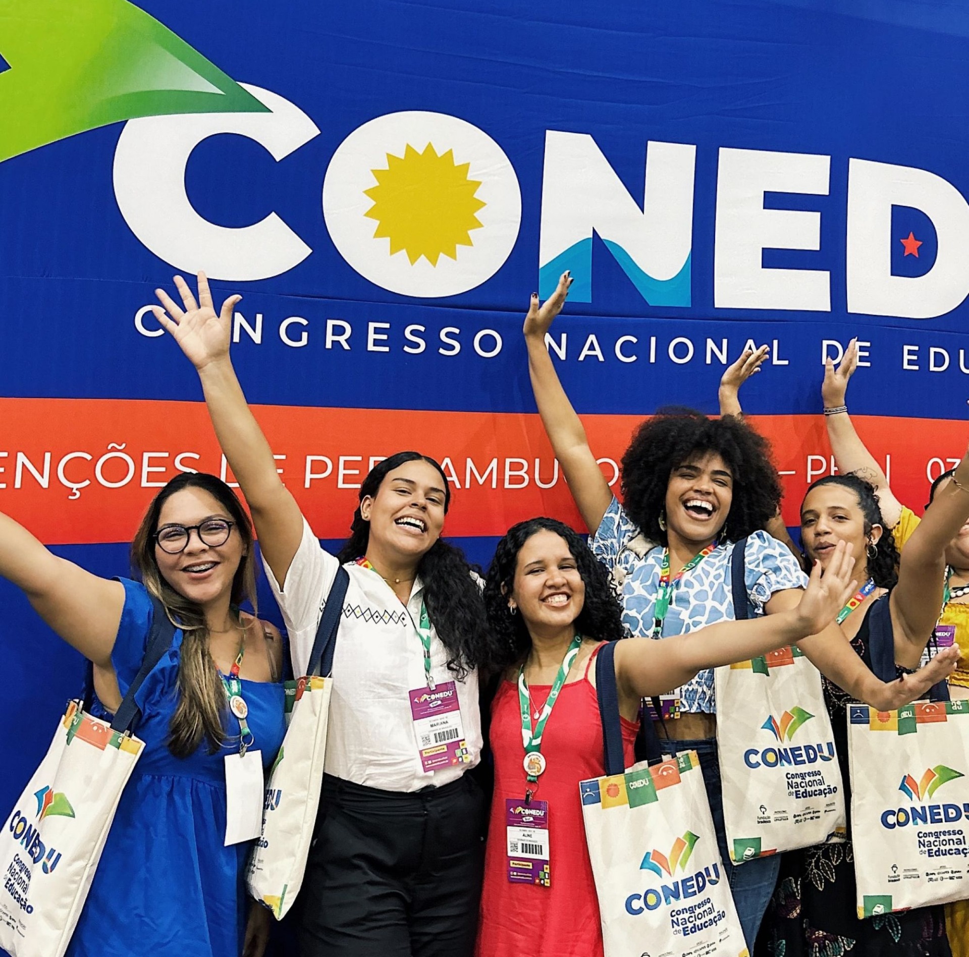 Cinco mulheres sorridentes acenam com os braços levantados em frente a um painel azul e vermelho do "CONEDU". Elas usam crachás no pescoço e carregam sacolas ecológicas brancas do evento com a logo colorida. A mulher ao centro veste vestido vermelho, e as demais usam blusas e vestidos nas cores azul, branca e estampada.