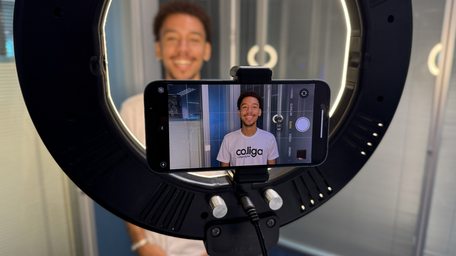 Fotografia colorida em plano médio e ângulo frontal, capturada através dos bastidores de uma gravação ou sessão fotográfica. O foco principal está na ecrã de um smartphone acoplado no centro de um iluminador circular (ring light).
