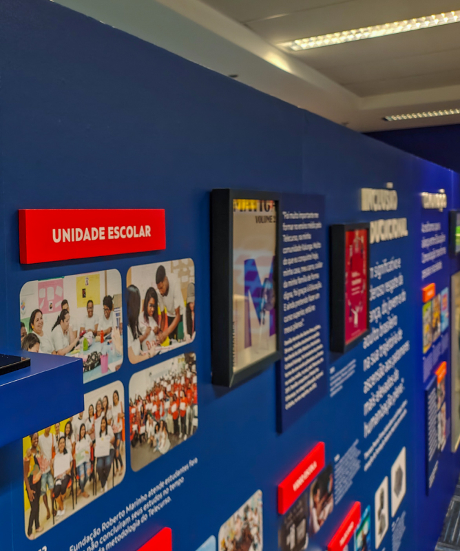 Vista de um painel expositivo com o troféu do Prêmio Jovem Cientista com o tema “Conectividade e Inclusão Digital”. A parede apresenta iniciativas educacionais da Fundação Roberto Marinho, sob o eixo “Unidade Escolar”, com fotos de estudantes e professores em atividades escolares e de tecnologia. Ao fundo, são visíveis outros segmentos temáticos como "Inclusão Vocacional" e "Trabalho".