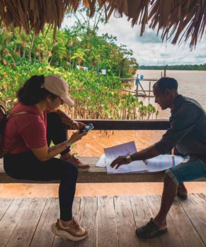 Dois adultos sentados sob um quiosque de madeira com cobertura de palha, à beira de um rio em área de floresta amazônica. À esquerda, uma mulher com boné claro, mochila vermelha e camiseta rosa segura um celular e observa um papel. À direita, um homem de bermuda jeans e camisa escura aponta para mapas ou documentos sobre o banco. O ambiente é ribeirinho, com vegetação densa e o rio ao fundo, transmitindo um clima de conversa e troca de saberes em meio à natureza.