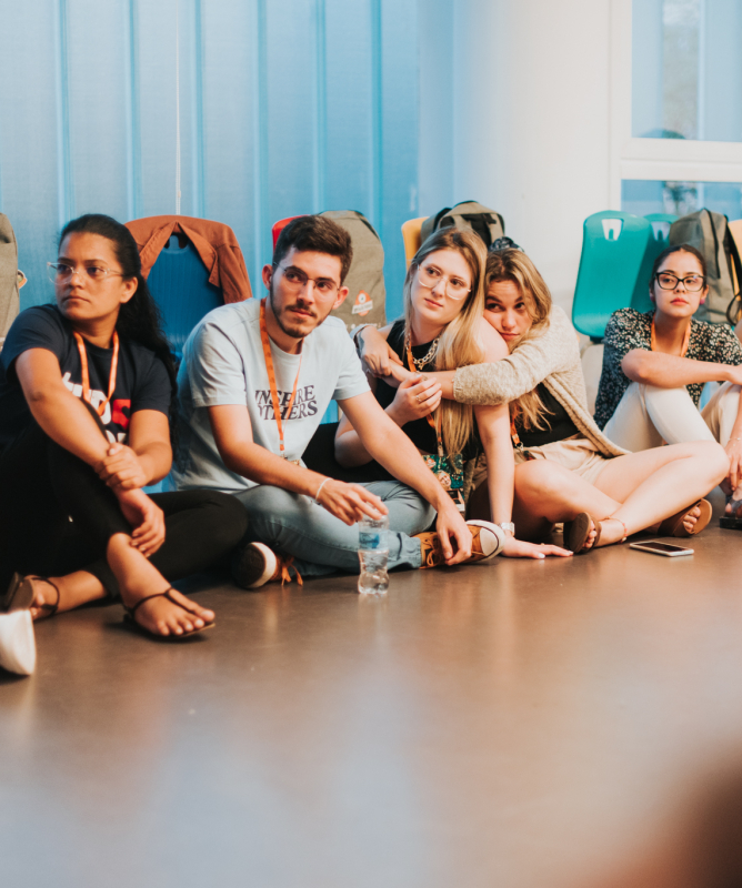 Um grupo de jovens está reunido em roda dentro de uma sala ampla, iluminada por luz natural. Todos estão sentados no chão ou em cadeiras, em clima de descontração e escuta. Ao centro, uma jovem fala enquanto os demais observam com atenção. O ambiente é acolhedor e transmite o sentimento de encerramento e afeto entre o grupo.
