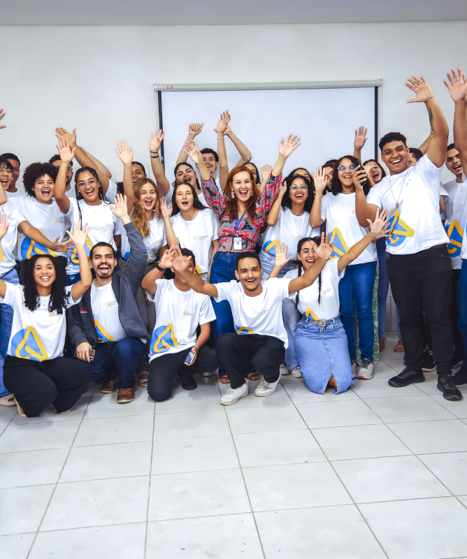 Um grupo de cerca de 20 jovens está reunido em uma sala de aula clara, com carteiras azuis ao redor. A maioria veste camisetas brancas com um logotipo azul e amarelo, e todos sorriem, levantando os braços para o alto em gesto de comemoração e alegria. No centro, uma mulher de camisa colorida também sorri com entusiasmo, acompanhando o grupo. O clima é de união, energia positiva e celebração coletiva.