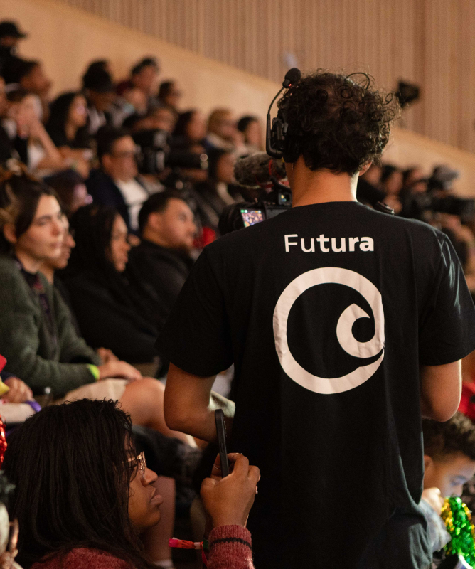 Plateia de um auditório lotado, com pessoas sentadas e atentas à programação. Em primeiro plano, de costas, está um cinegrafista usando fones de ouvido e camiseta preta com o logotipo do Canal Futura, que grava o evento. Algumas pessoas do público seguram celulares e objetos coloridos nas mãos. O ambiente tem iluminação suave e paredes revestidas de madeira clara.