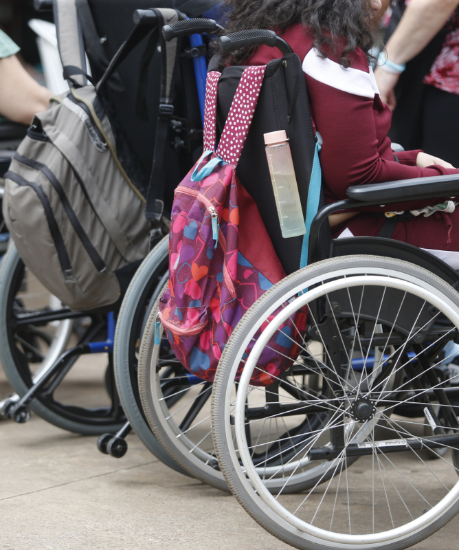 Uma foto que foca nas rodas e nos encostos de três cadeiras de rodas em um espaço externo. Duas pessoas estão sentadas nas cadeiras, mas apenas a parte de baixo de seus corpos aparece. Uma mochila colorida, com bolinhas, está pendurada no encosto da cadeira de rodas em primeiro plano.