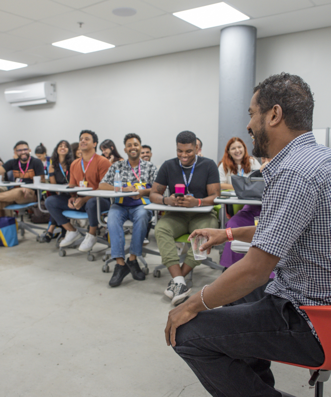 Na imagem, um grupo de jovens universitários está reunido em uma sala de aula moderna. Eles estão sentados em cadeiras com rodinhas e pequenas mesas acopladas, formando um semicírculo. Todos prestam atenção e sorriem para um homem que está à frente, sentado em uma cadeira vermelha, conversando de forma descontraída. A sala é clara, com paredes brancas, ar-condicionado e iluminação de teto. O clima é leve e de troca, com os estudantes demonstrando interesse e engajamento na atividade.
