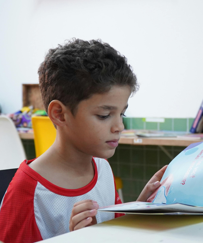 A imagem mostra uma criança em uma escola, sentada em uma carteira escolar e segurando e lendo um livro didático com as mãos. 