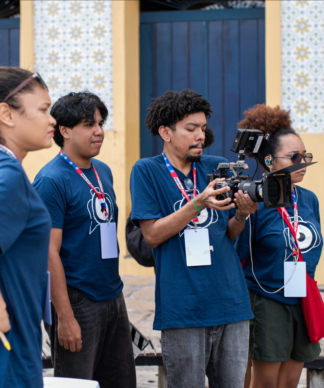 Um grupo de jovens está reunido em uma rua de paralelepípedos, em frente a uma parede amarela decorada com azulejos brancos e azuis. Todos vestem camisetas azul-marinho com crachás vermelhos pendurados no pescoço. No centro, um rapaz segura uma câmera de cinema, focado na filmagem, enquanto os colegas observam atentamente. A atmosfera é de aprendizado e trabalho em equipe durante uma atividade prática de gravação.