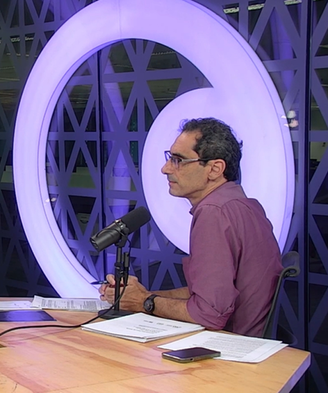 Uma mulher branca, de cabelos curtos, e um homem branco, de cabelos castanhos, estão sentados em uma mesa a frente de microfones. O cenário é bastante iluminado e há um cenário oluminado atrás formando a logo do Canal Futura. Há papeis sobre a mesa e o clima é de conversa e concentração.