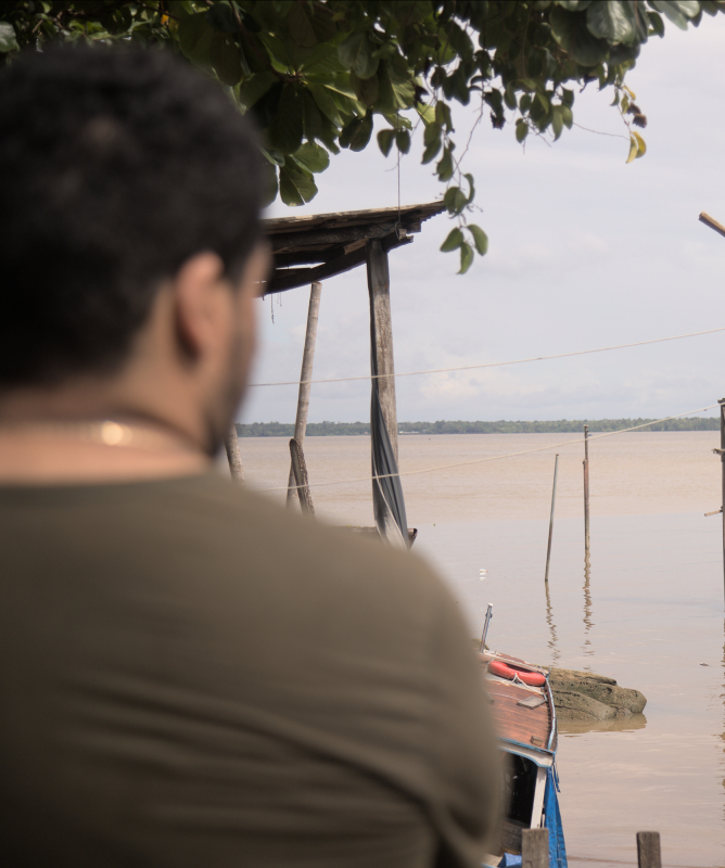 À beira de um rio de águas amarronzadas, casas de madeira sobre palafitas se estendem sobre o espelho d’água. Um homem observa a paisagem, parcialmente de costas. Pequenos barcos coloridos estão atracados e o céu nublado reforça o clima amazônico e ribeirinho.